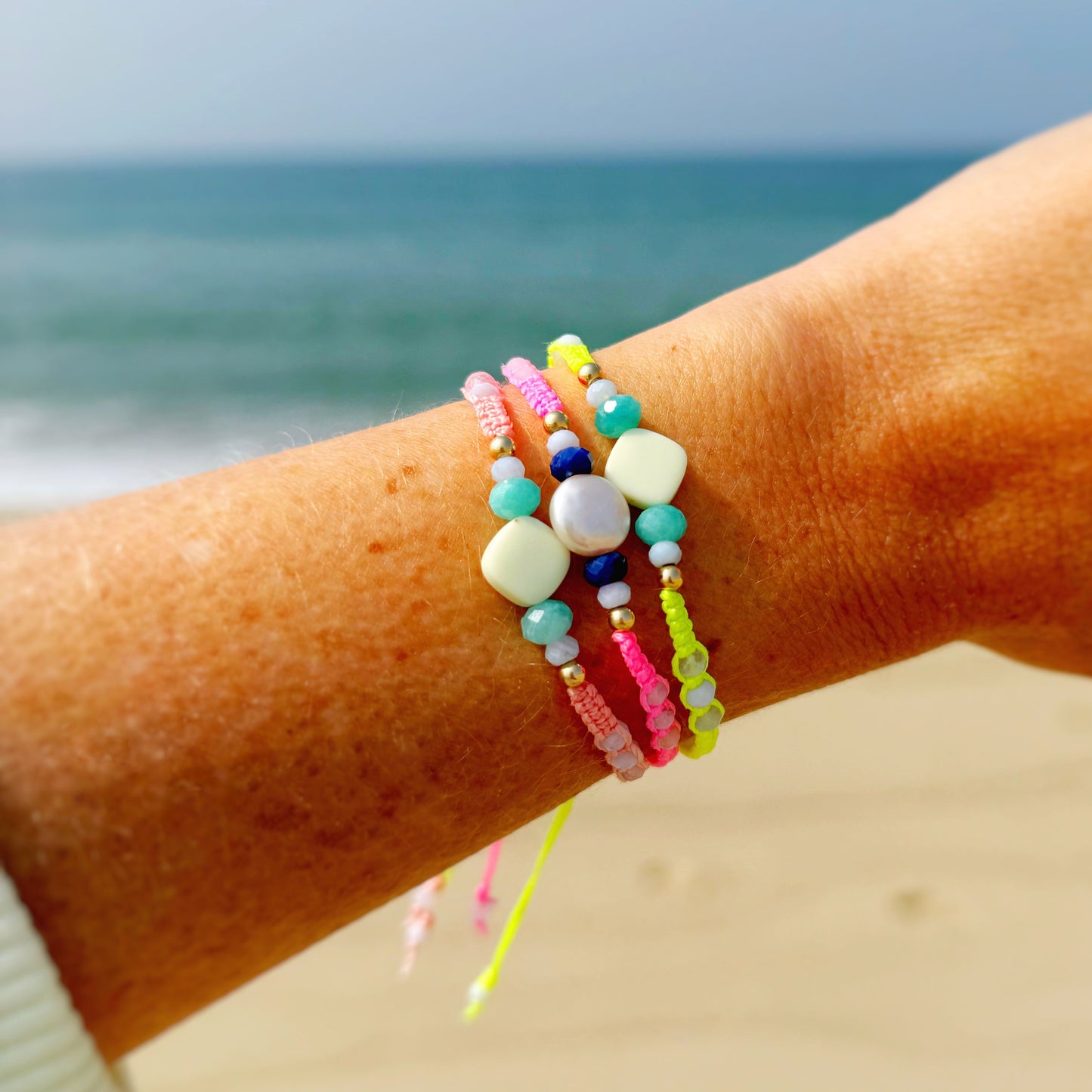 mermaids and madeleines macrame bracelets are created with cord, semiprecious beads and pearls. Friendship style bracelets with slide clasps. This is a photograph of 3 macrame bracelets on a wrist with a beach in a background