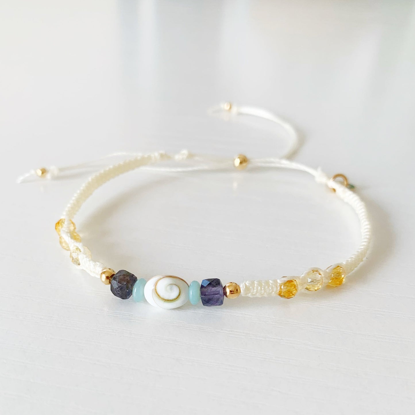 the mermaid money macrame bracelet by mermaids and madeleines features vanilla color cord, knotted with a shiva shell at the center, with amazonite, iolite and citrine beads, complimented with 14k gold filled beads and a slider. This bracelet is an adjustable friendship style bracelet and photographed on a white surface