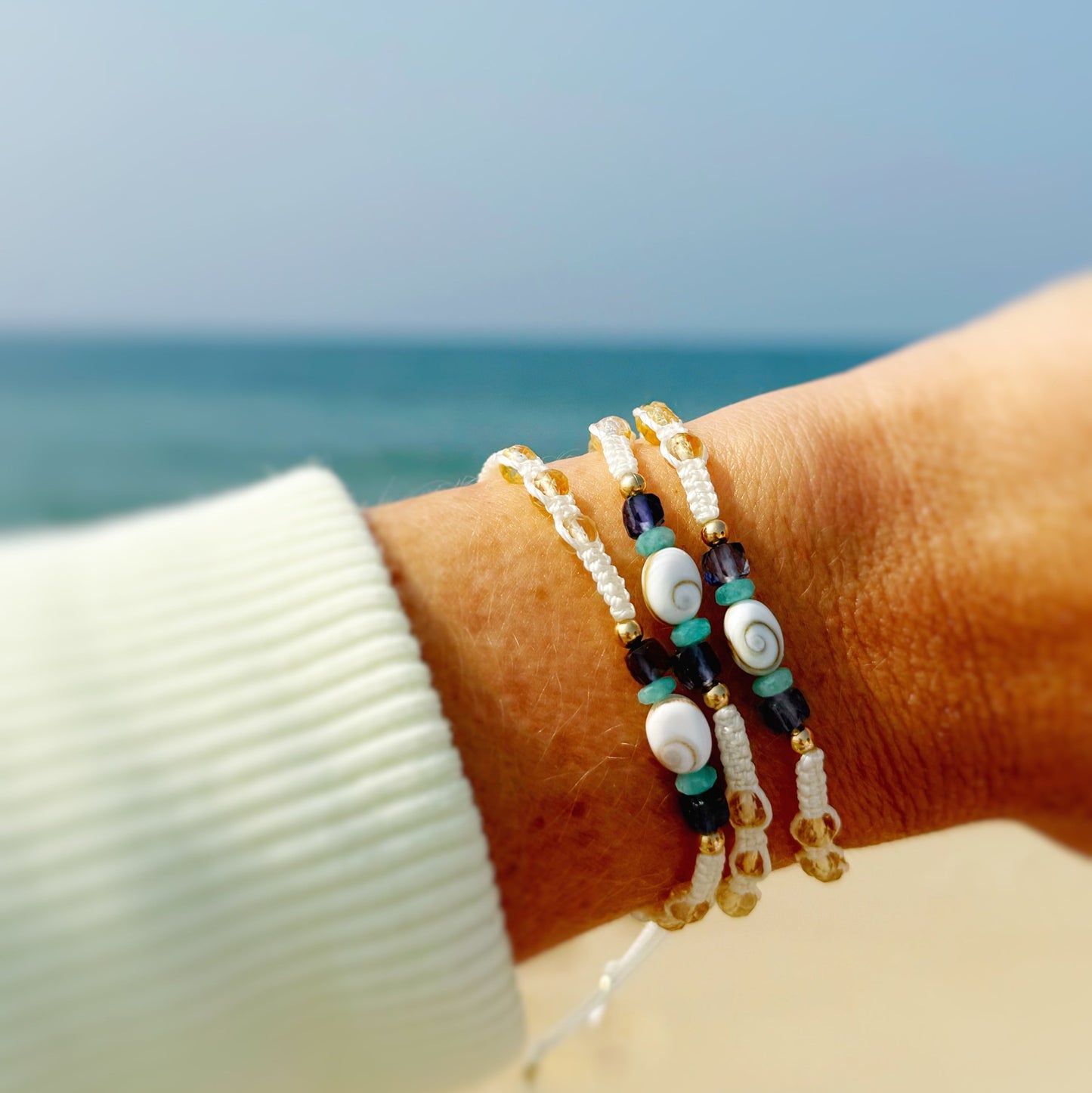 mermaid money macrame bracelets are created with vanilla color cord, macrame knotted into an adjustable friendship bracelet with a shiva shell at the center of the bracelet complimented by abundance semiprecious beads including amazonite, iolite and citrine, with 14k gold filled beads and 14k gold filled slide clasps. This set of 3 macrame bracelets is photographed as a stack on a wrist with a beach in the background