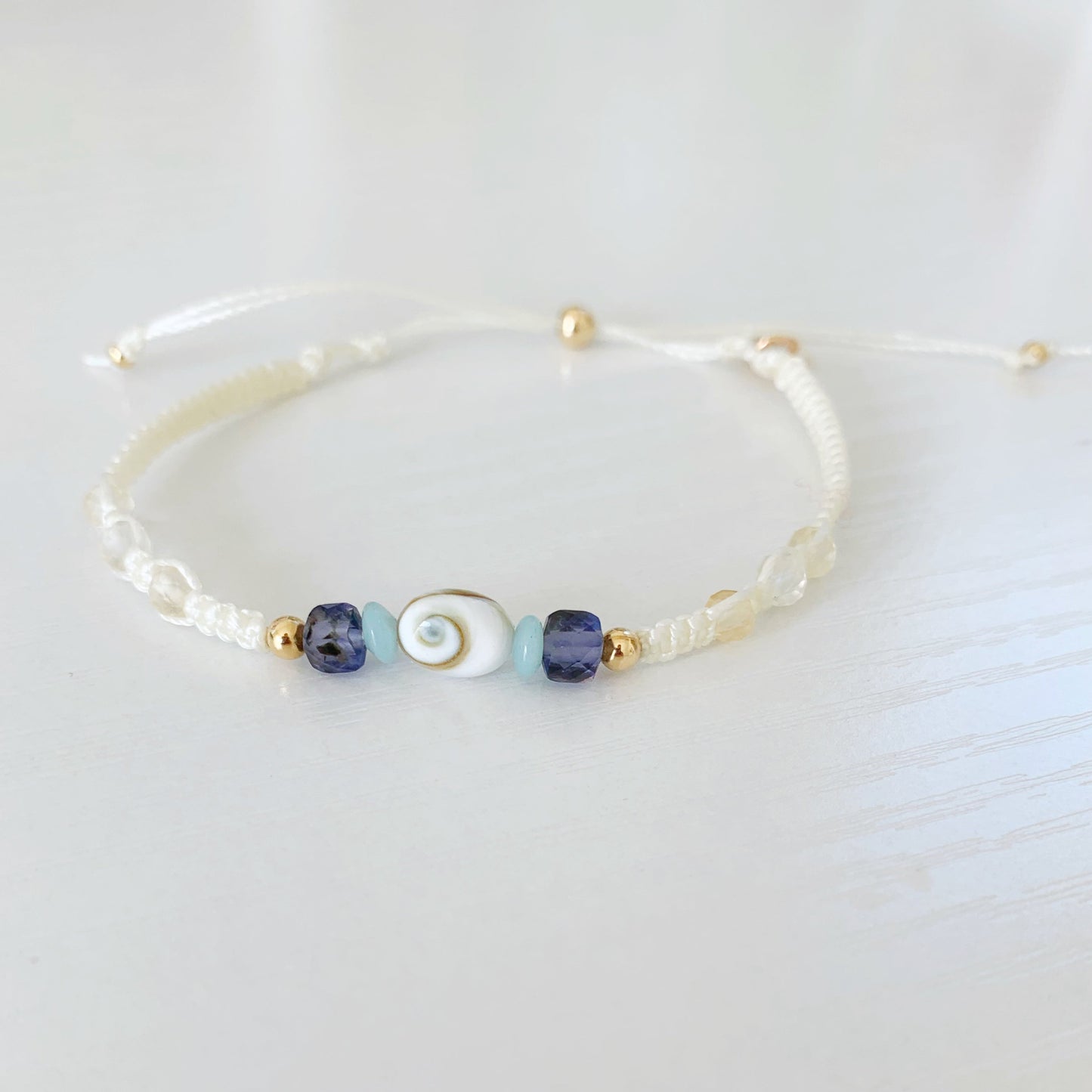 The mermaid money macrame bracelet by mermaids and madeleines is a vanilla cord bracelet created in an adjustable friendship bracelet style. The bracelet has a shiva shell at the center with teal amazonite, blue/purple iolite and soft citrine beads with 14k gold filled beads and a slider clasp. this bracelet is photographed on a white surface
