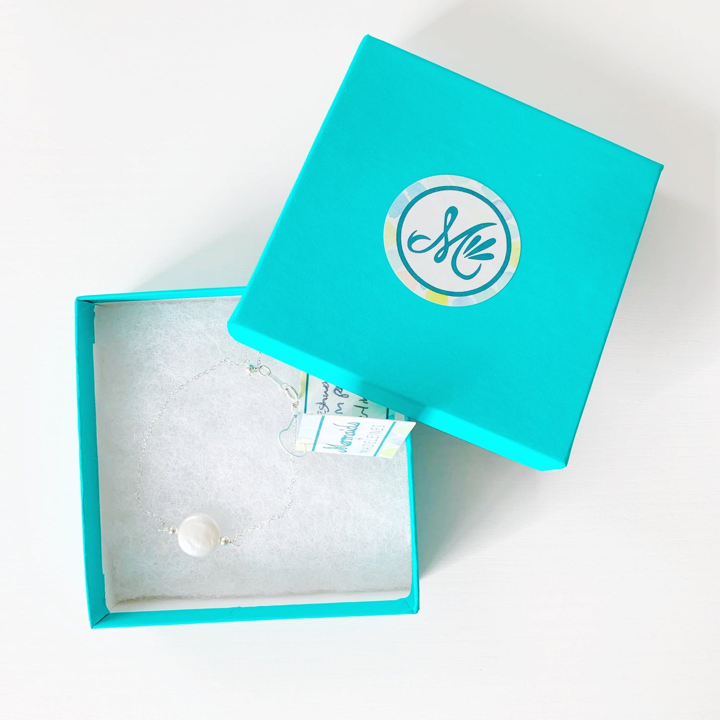 the newport bracelet by mermaids and madeleines is a sterling silver chain bracelet with a freshwater coin pearl at the center and with a slide bead near the clasp to make it adjustable. this bracelet is photographed in a teal gift box on a white surface