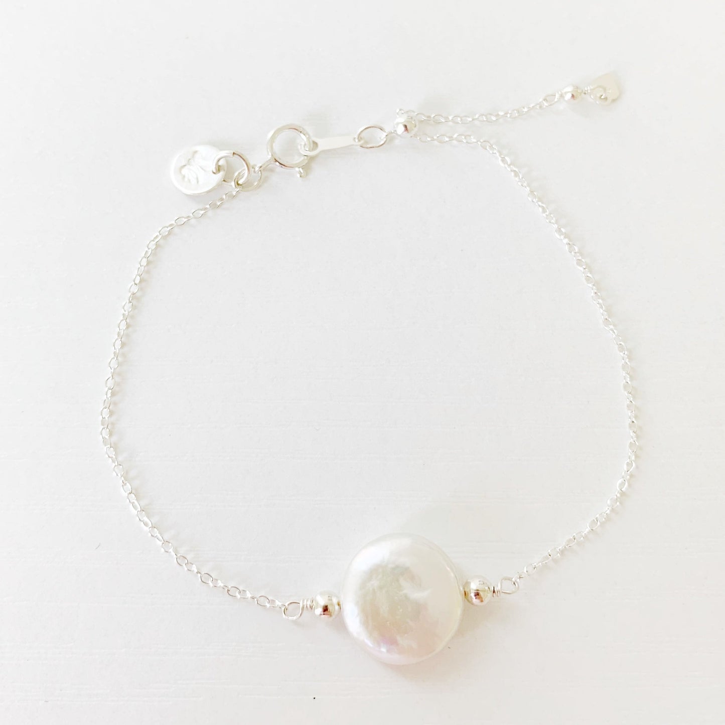 this bracelet has a freshwater coin pearl at the center on sterling silver chain with a slide bead near the clasp to adjust the length. this bracelet is photographed from a top down view on a white surface