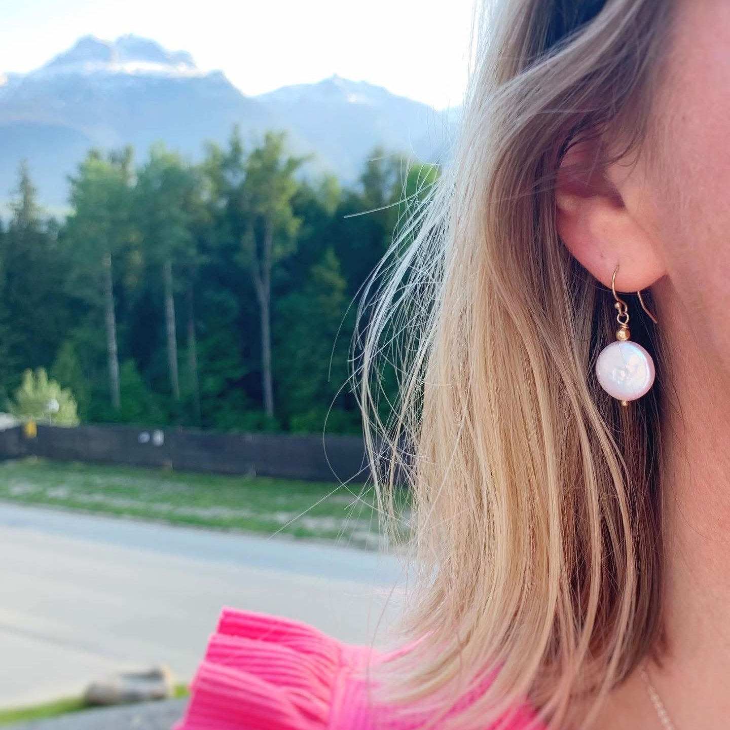Person wearing a pink top and coin pearl drop earring with a scenic background of trees and mountains.