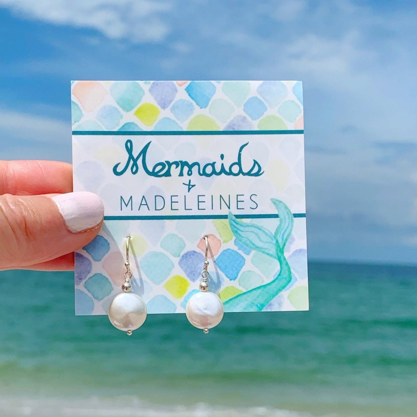 Newport pearl earrings are created with freshwater coin pearls and sterling silver findings. this pair is pictured on a mermaids and madeleines earring card and photographed being held up with a beach background