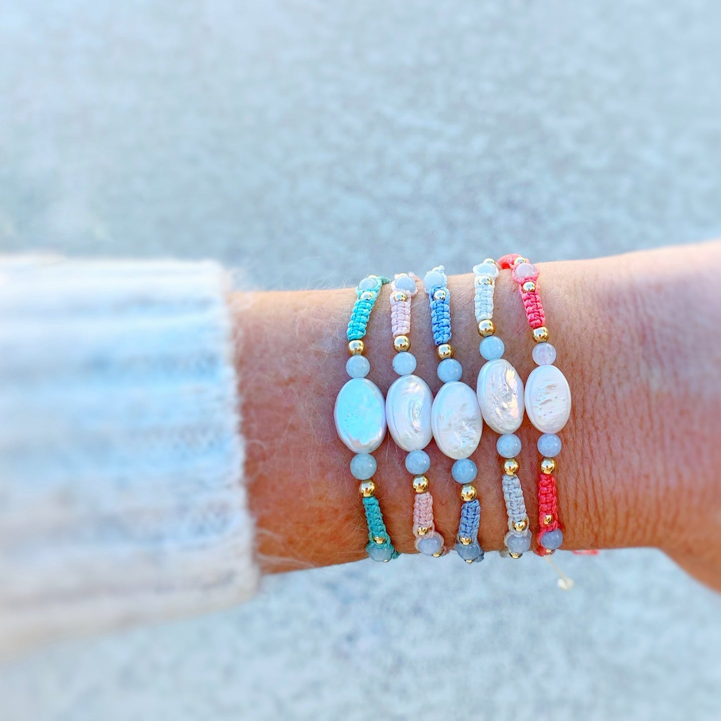the orleans bracelets by mermaids and madeleines are created with macrame cord with freshwater oval pearls at the center and complimented by round aquamarine beads and 14k gold filled beads. this is a photograph of a wrist with a little bit of sweater showing and all 5 orleans bracelet color options are on the wrist.