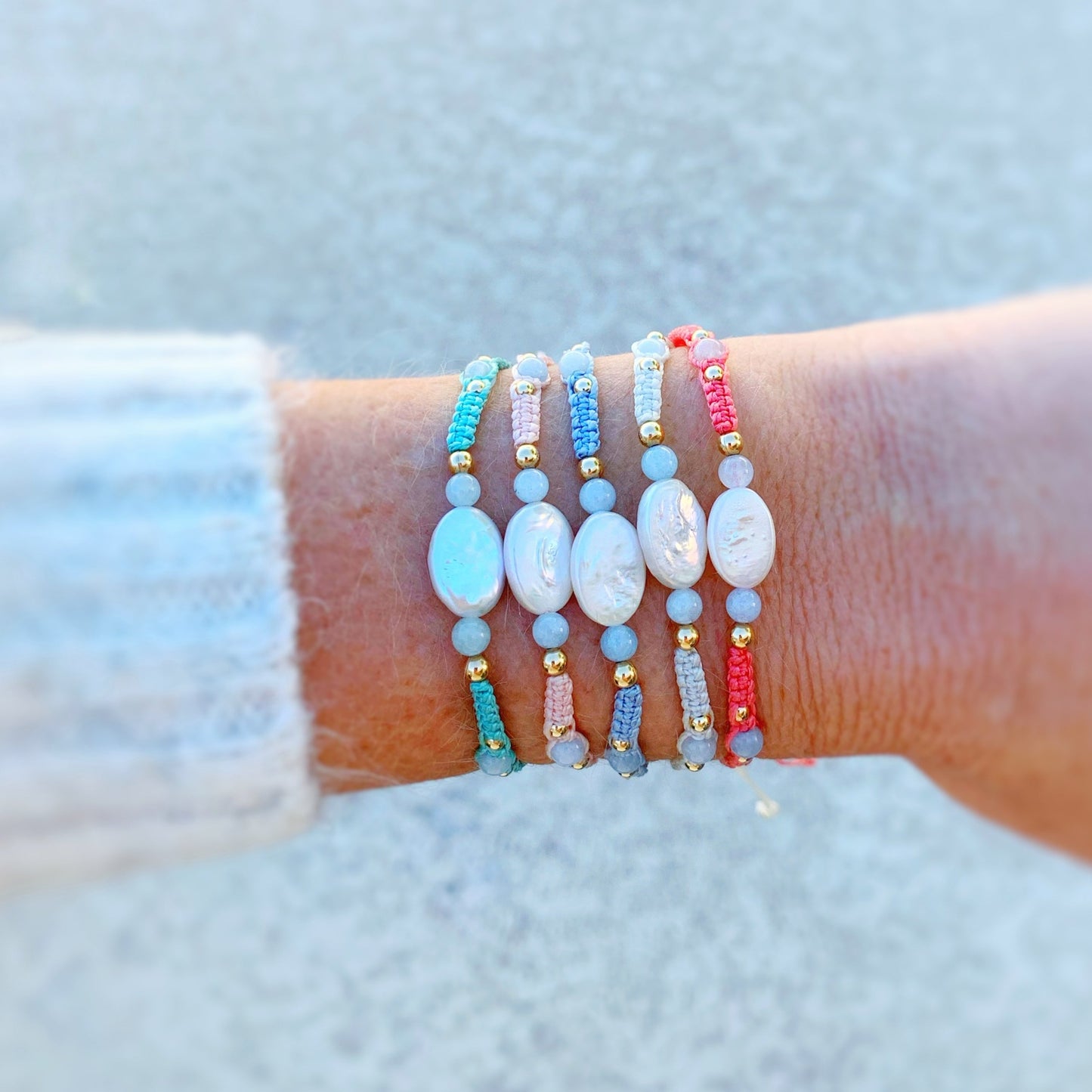 the orleans bracelets by mermaids and madeleines are created with macrame cord with freshwater oval pearls at the center and complimented by round aquamarine beads and 14k gold filled beads. this is a photograph of a wrist with a little bit of sweater showing and all 5 orleans bracelet color options are on the wrist.
