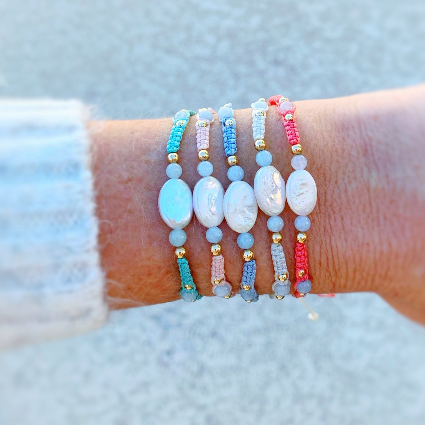 the orleans bracelets by mermaids and madeleines are created with macrame cord with freshwater oval pearls at the center and complimented by round aquamarine beads and 14k gold filled beads. this is a photograph of a wrist with a little bit of sweater showing and all 5 orleans bracelet color options are on the wrist.