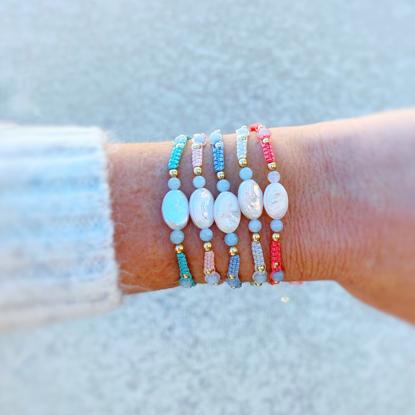 the orleans bracelets by mermaids and madeleines are created with macrame cord with freshwater oval pearls at the center and complimented by round aquamarine beads and 14k gold filled beads. this is a photograph of a wrist with a little bit of sweater showing and all 5 orleans bracelet color options are on the wrist.