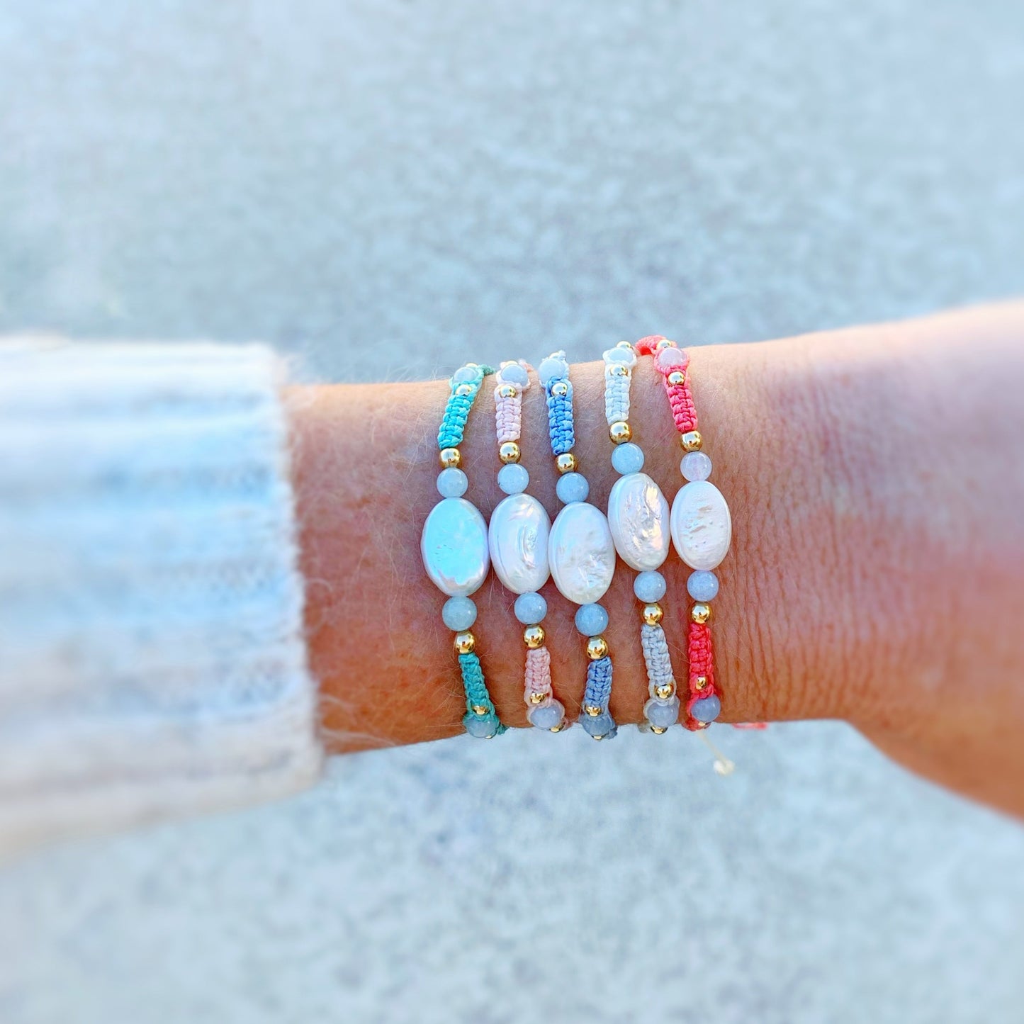 the orleans bracelets by mermaids and madeleines are created with macrame cord with freshwater oval pearls at the center and complimented by round aquamarine beads and 14k gold filled beads. this is a photograph of a wrist with a little bit of sweater showing and all 5 orleans bracelet color options are on the wrist.
