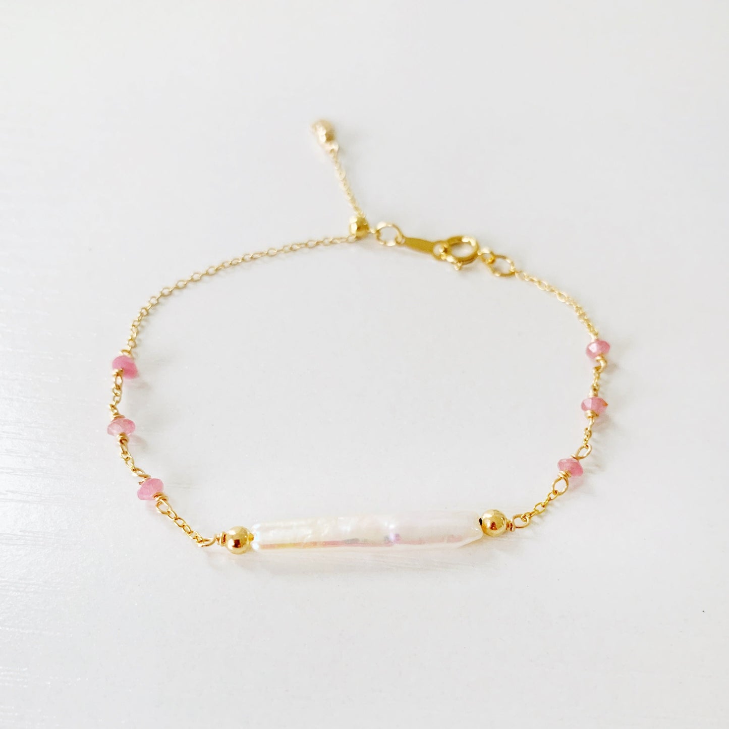 the pink azalea adjustable bracelet by mermaid and madeleines in a 14k gold filled chain bracelet with a slide bead at the clasp. the bracelet features a freshwater pearl bar at the center with pink tourmaline beads going down the chain. this bracelet is photographed flat on a white surface with a front view