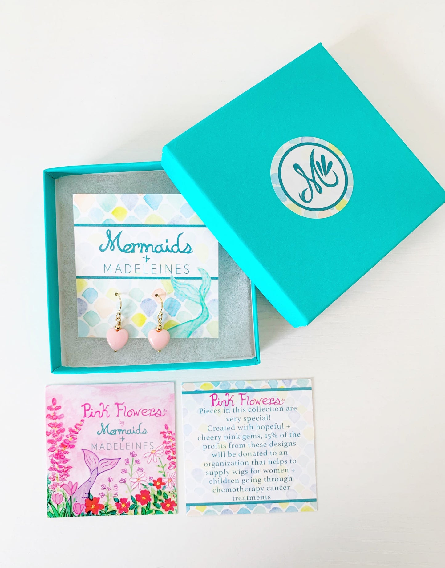 the pink posy earrings by mermaids and madeleines are light pink queen conch shell beads carved into hearts and topped with 14k gold filled round bead and feature 14k gold filled earring wires. this pair is carded and placed in a teal gift box with the pink flowers story card on the table in front of the box. all items are photographed on a white surface