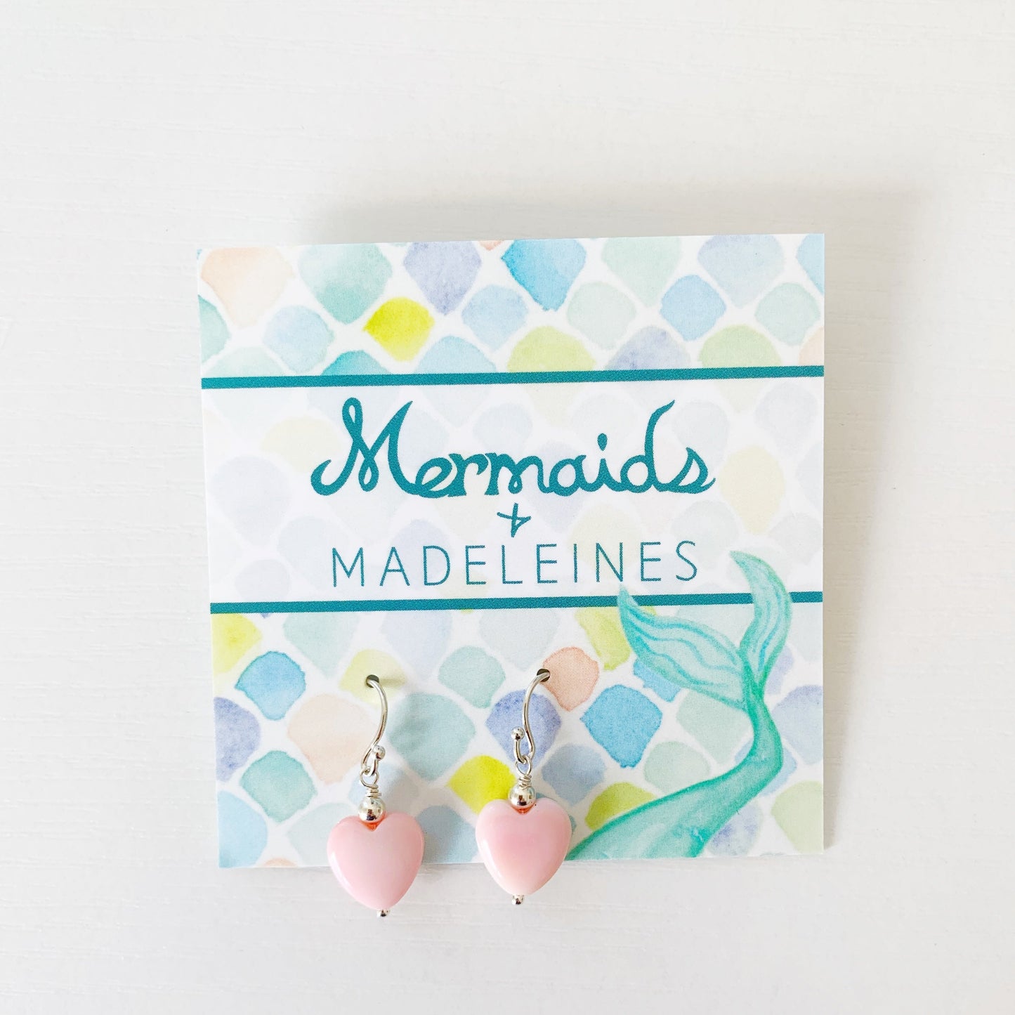 The pink posy earrings by mermaids and madeleines are light pink queen conch shell carved hearts with sterling silver beads and findings. this pair is carded and photographed on a white surface