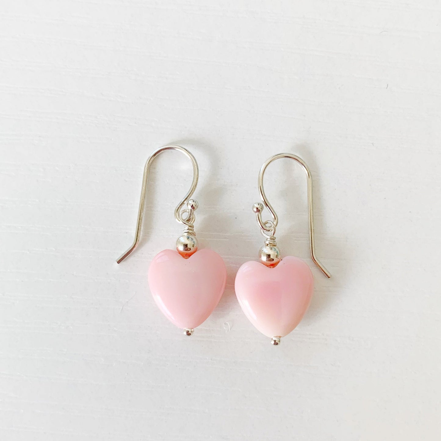 the pink posy earrings by mermaids and madeleines are light pink queen conch shell beads carved into hearts. Each has a sterling silver bead on top and features sterling silver findings. this pair of earrings is photographed on a white surface