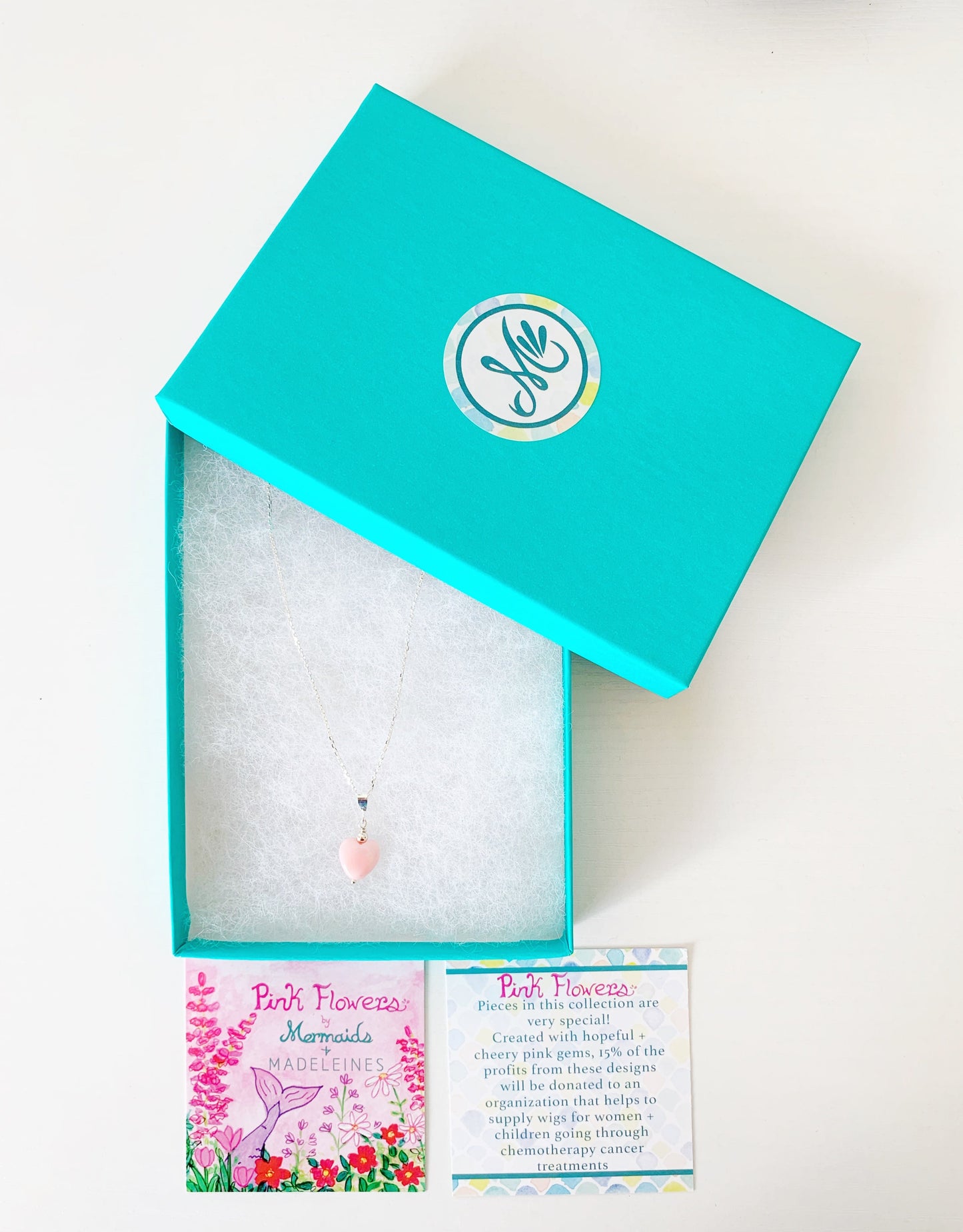the pink posy necklace by mermaids and madeleines is a pendant style necklace with a light pink queen conch shell carved into a heart shaped bead. Theres a small sterliing silver bead on top of this drop and the necklace features sterling silver findings and chain. This necklace is placed in a teal gift box with a pink flower collection story card set in front of the box. all items are photographed on a white surface