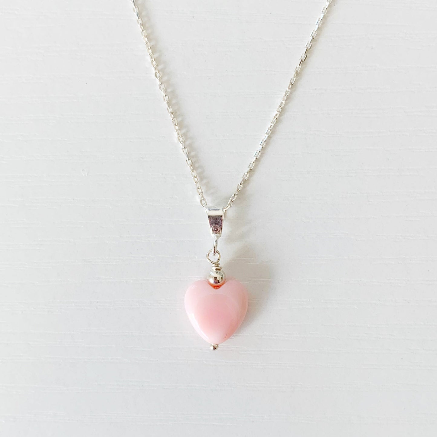 the pink posy necklace by mermaids and madeleines is a pendant style necklace with a light pink queen conch shell bead carved into a heart shape. theres a small sterling silver round bead on top of this drop. The necklace has sterling silver findings and chain. This necklace is photographed close up on a white surface