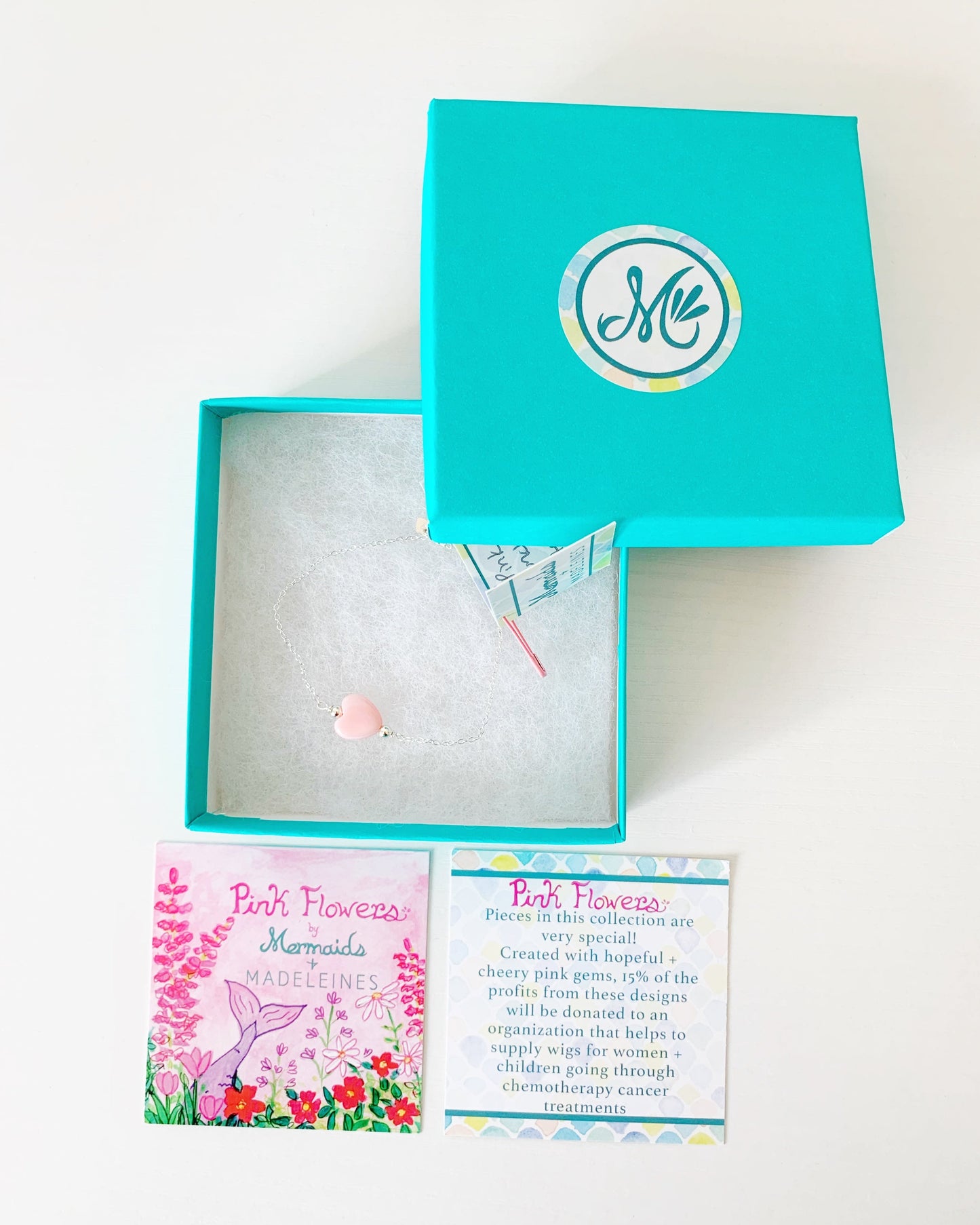 the pink posy adjustable bracelet by mermaids and madeleines is a chain style bracelet with a pink conch shell heart bead at the center and a slide bead near the clasp to make it adjustable. this bracelet is photographed in a teal gift box with a pink flower story card below it all on a white surface