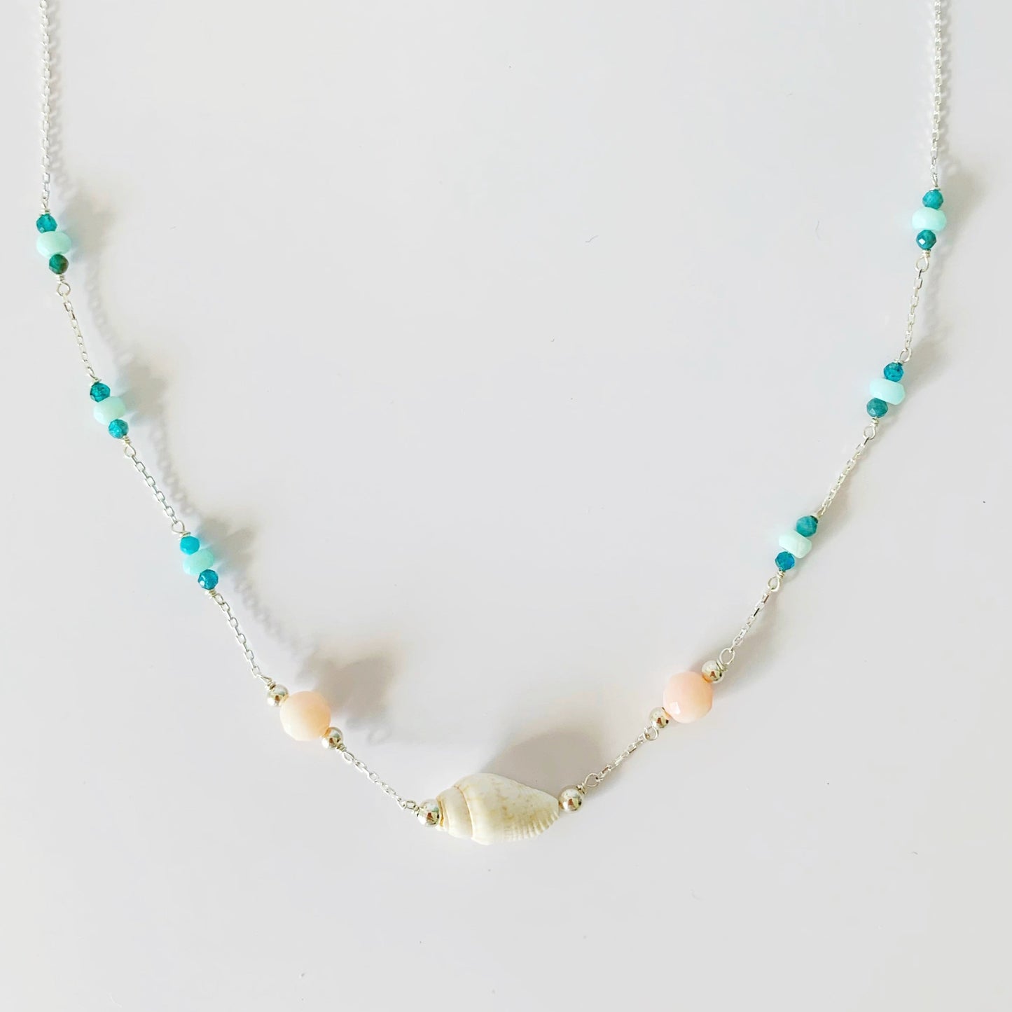 the pool party shellebration necklace by mermaids and madeleines is station style necklace with a shell and coral beads at the center and little sections of Peruvian opal and apatite throughout the sterling chain to the clasp. this necklace is photographed on a white surface