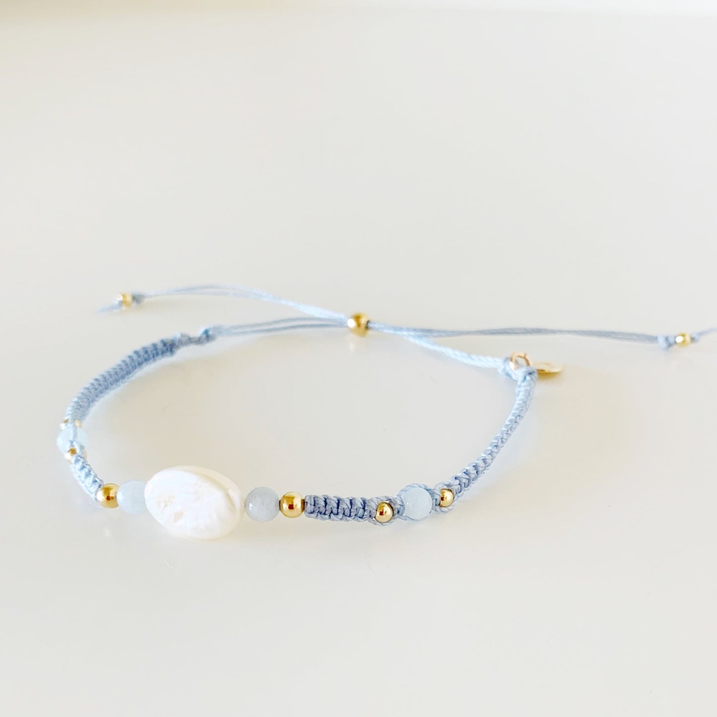 the regal seagull bracelet by mermaids and madeleines is an adjustable macrame bracelet with blue-gray cord with a freshwater oval pearl at the center with 4mm aquamarine beads on either side. this bracelet is photographed from a slight right angle with the piece on a white surface