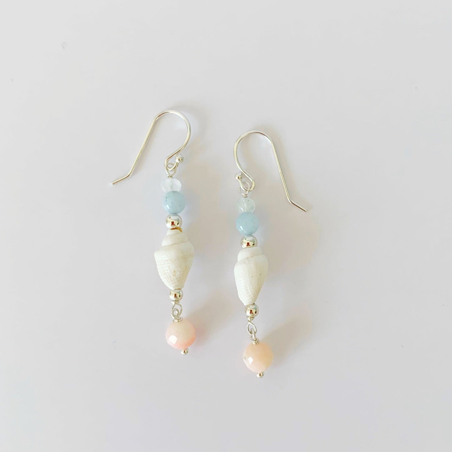 the seersucker shellebration earrings by mermaids and madeleines are a linear drop style earrings with a shell at the center complimented by moonstone, aquamarine, and with a coral bead drop at the bottom. this pair is photographed on a white surface