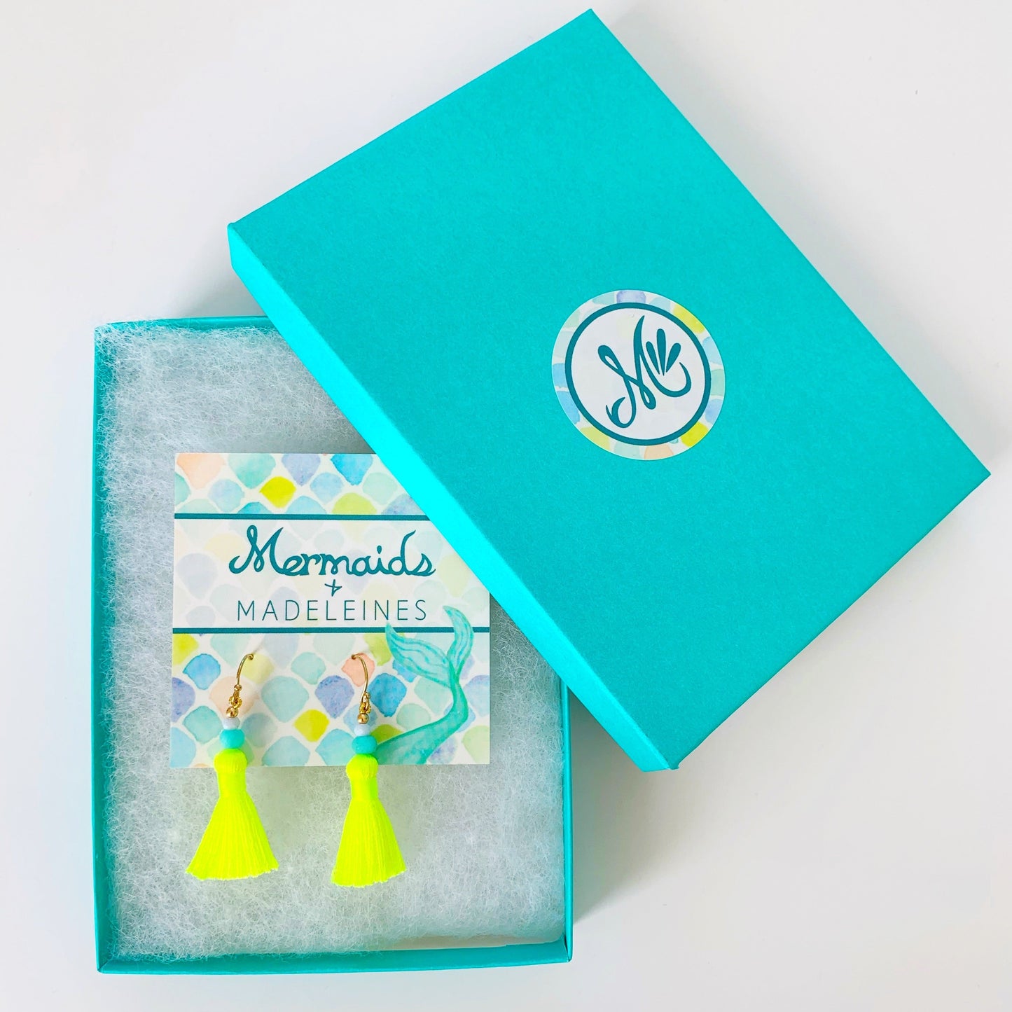 Turquoise box with 'Mermaids + Madeleines' branding, containing neon yellow tassel earrings on an earring card.