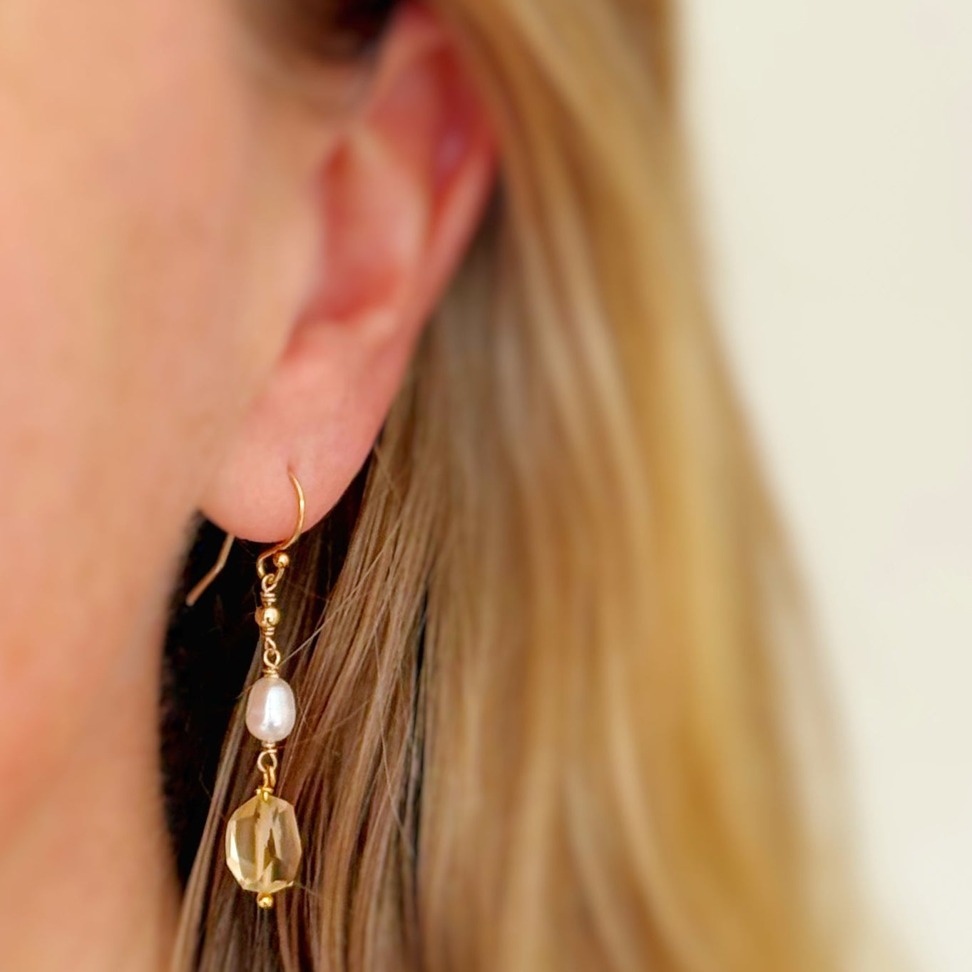 a close up of a person wearing a gold dangle earring that has a small freshwater pearl and nugget shaped faceted citrine drop.