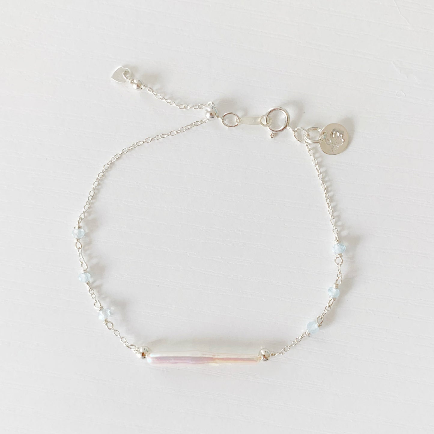 the truro bracelet by mermaids and madeleines is an adjustable chain style bracelet with a slide bead at the clasp. The bracelet features a freshwater stick pearl at the center with small faceted aquamarine beads in stations on either side. the findings, slide clasp and chain are all sterling silver. this bracelet is photographed in a top down view on a white surface
