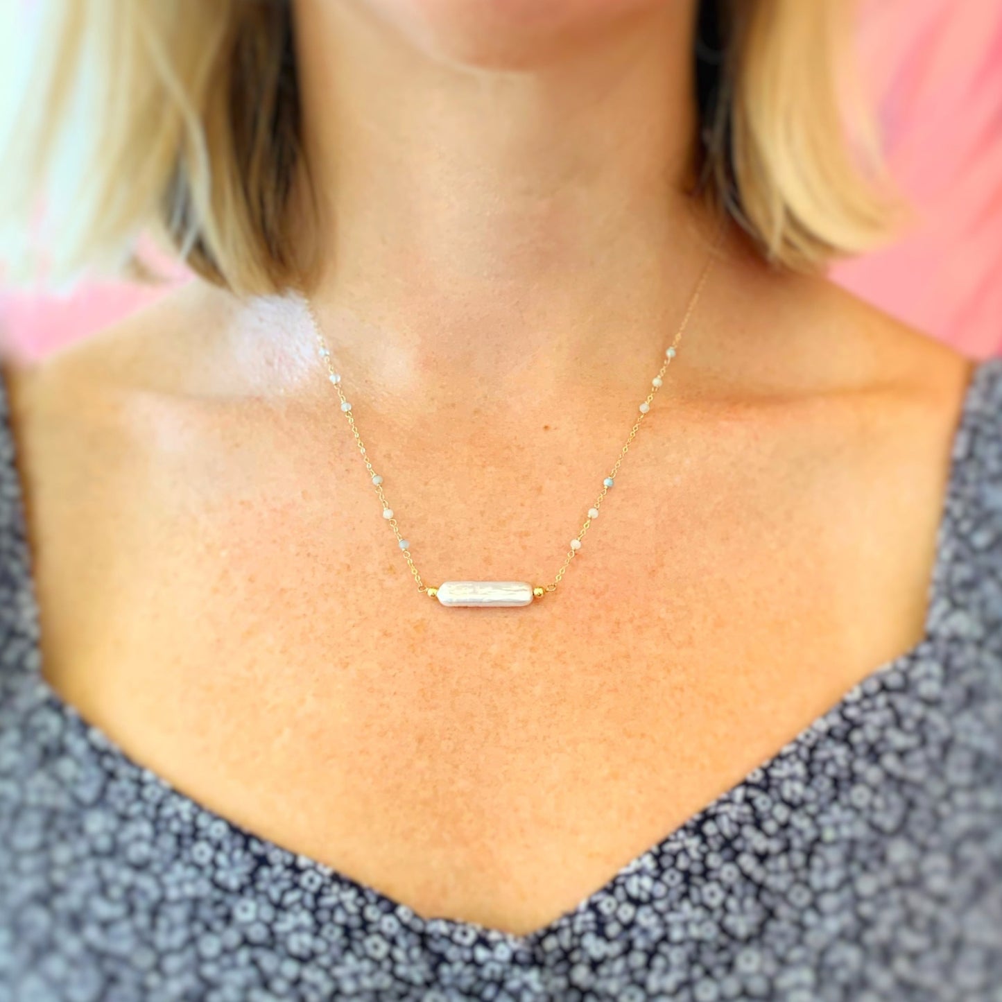 the truro necklace by mermaids and madeleines is available in 14k gold filled or sterling silver metals. the necklace has a freshwater stick pearl at the center going from east to west and has microfacteted aqumarine beads dotting up the sides of the chain. the necklace photographed is 14k gold filled and pictured worn on a women with a closer up view. you can see a little bit of her shoulder length hair and her navy and white flowered shirt.