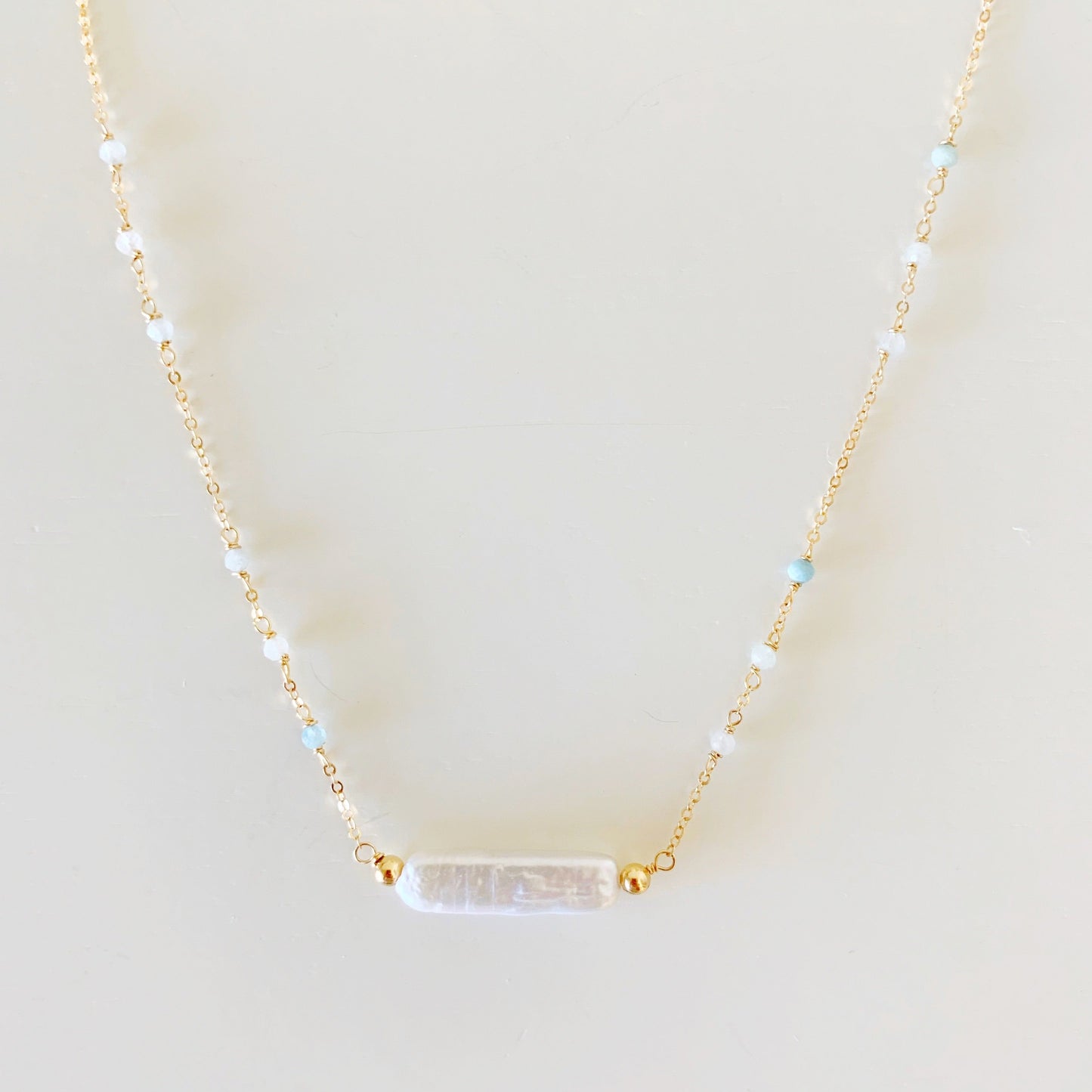 The truro necklace is designed with 14k gold filled chain and findings and a freshwater stick pearl at the center with microfaceted aquamarine along the chain up the necklace. this necklace is photographed closer up on a white surface