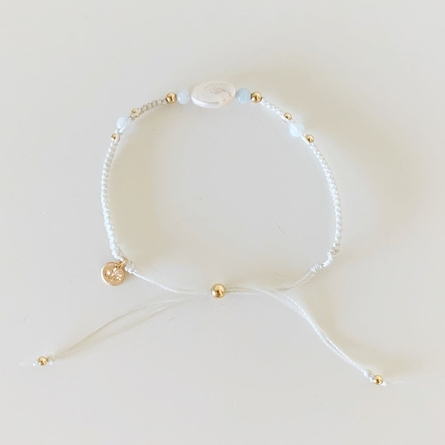 the world is your oyster bracelet by mermaids and madeleines is an adjustable macrame bracelet designed with white shell cord and freshwater oval pearl at the center with aquamarine beads on both sides. this bracelet is photographed from the top town in full view on a white surfaace