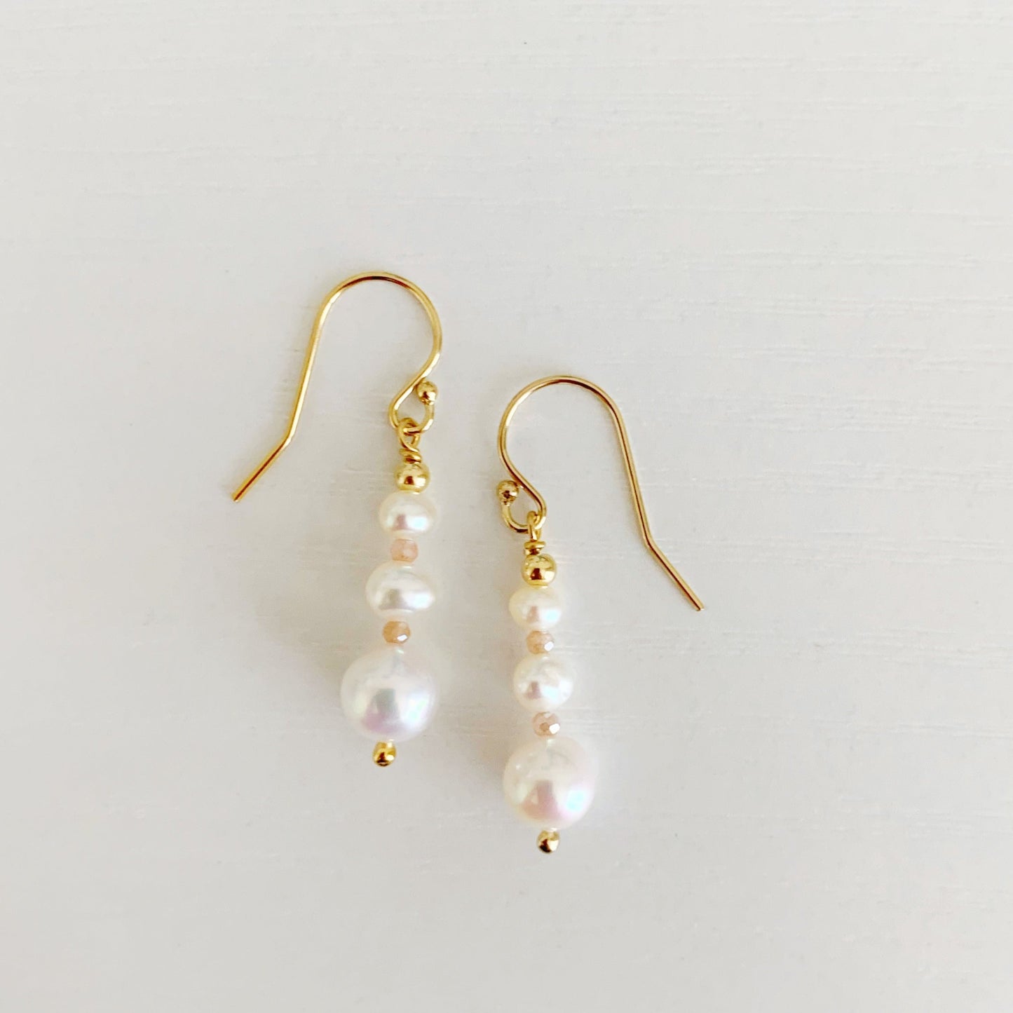 Barrington earrings by mermaids and madeleines feature freshwater pearls in a linear taper, with peach moonstone accents and 14k gold filled findings. this pair is photographed flat on a white surface with one earring placed slightly higher than the other on the surface