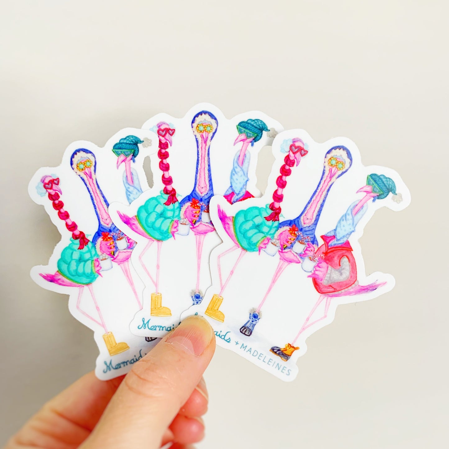 this is a photograph of 3 vinyl stickers held up and fanned out in a hand with a white background. the stickers are illustrations of 3 flamingos dressed in winter wear and designed by mermaids and madeleines