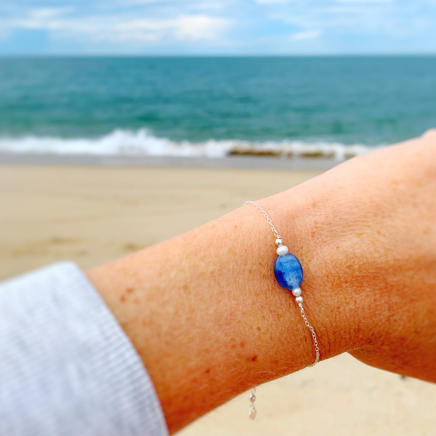 the brewster bracelet by mermaids and madeleines is an adjustable sterling silver chain bracelet with a slide bead at the clasp. there's a blue kyanite coin bead at the center complimented by small freshwater pearls on each side. this bracelet is photographed close up on a wrist with a beach background