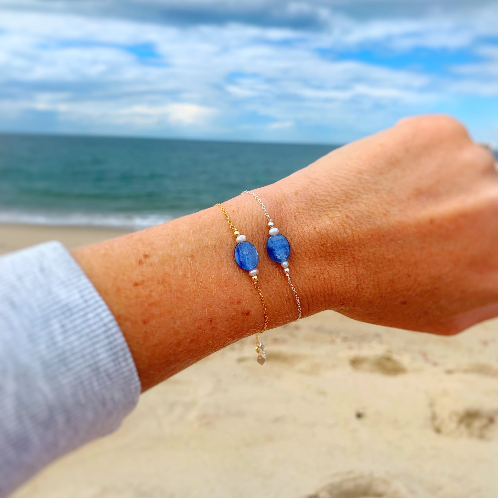 the brewster bracelet by mermaids and madeleines is an adjustable sterling silver chain bracelet with a slide bead at the clasp. there's a blue kyanite coin bead at the center complimented by small freshwater pearls on each side. this is a photo of the brewster bracelet in sterling silver and in 14k gold filled close up on a wrist with a beach background