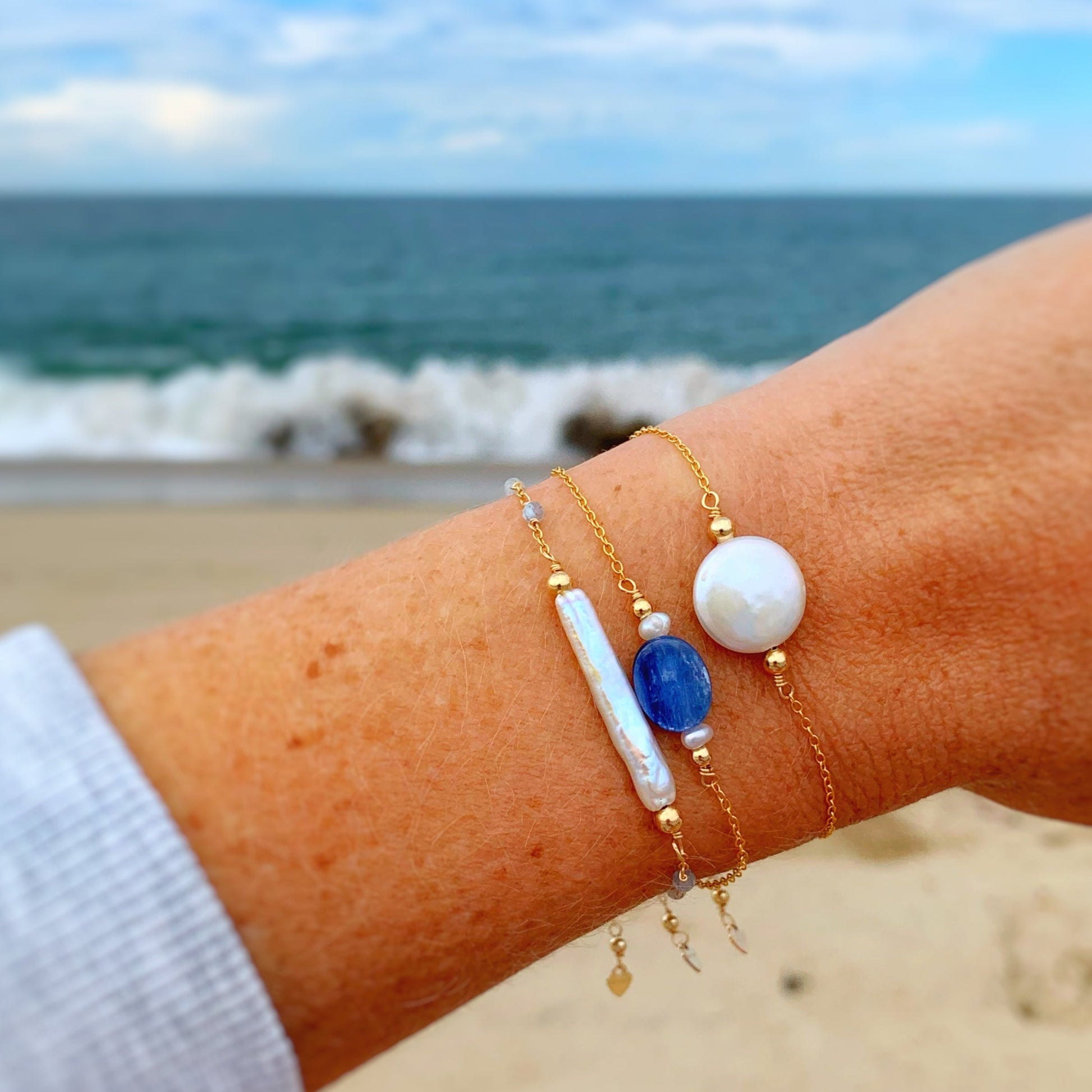 the brewster bracelet by mermaids and madeleines is an adjustable 14k gold filled chain bracelet with a slide bead at the clasp. there's a blue kyanite coin bead at the center complimented by small freshwater pearls on each side. this is a photo of the brewster bracelet, the truro bracelet and the newport bracelet close up on a wist with a beachy background