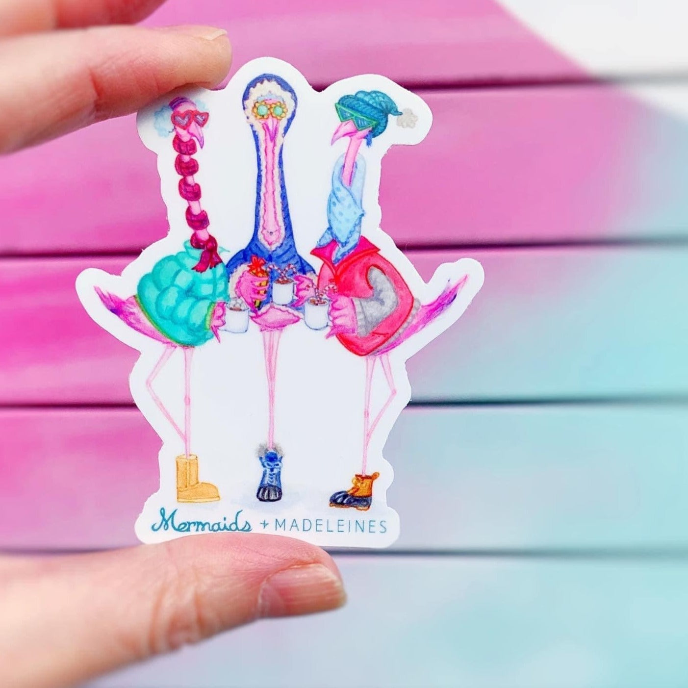 holiday flamingal vinyl sticker by mermaids and madeleines. this is a photograph of a vinyl sticker of 3 flamingos dressed in winter clothing illustrated by Heather Auclair Welch. The sticker is held up in a hand with a pink and aqua color backdrop