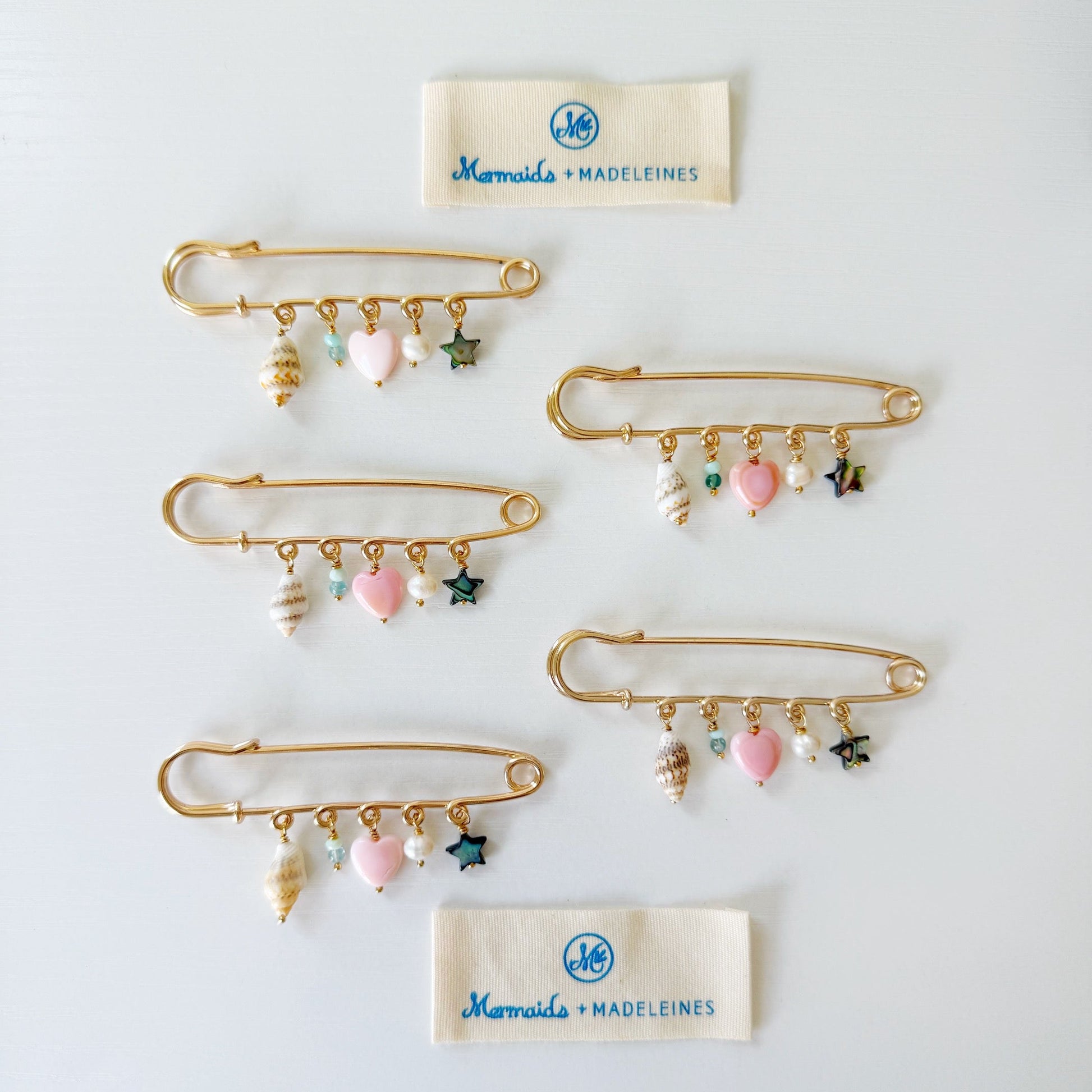 Six large, gold safety-pins with dangling seashell, pink heart, and star charms, arranged on a white background with two cream fabric labels reading Mermaid + Madeleine.