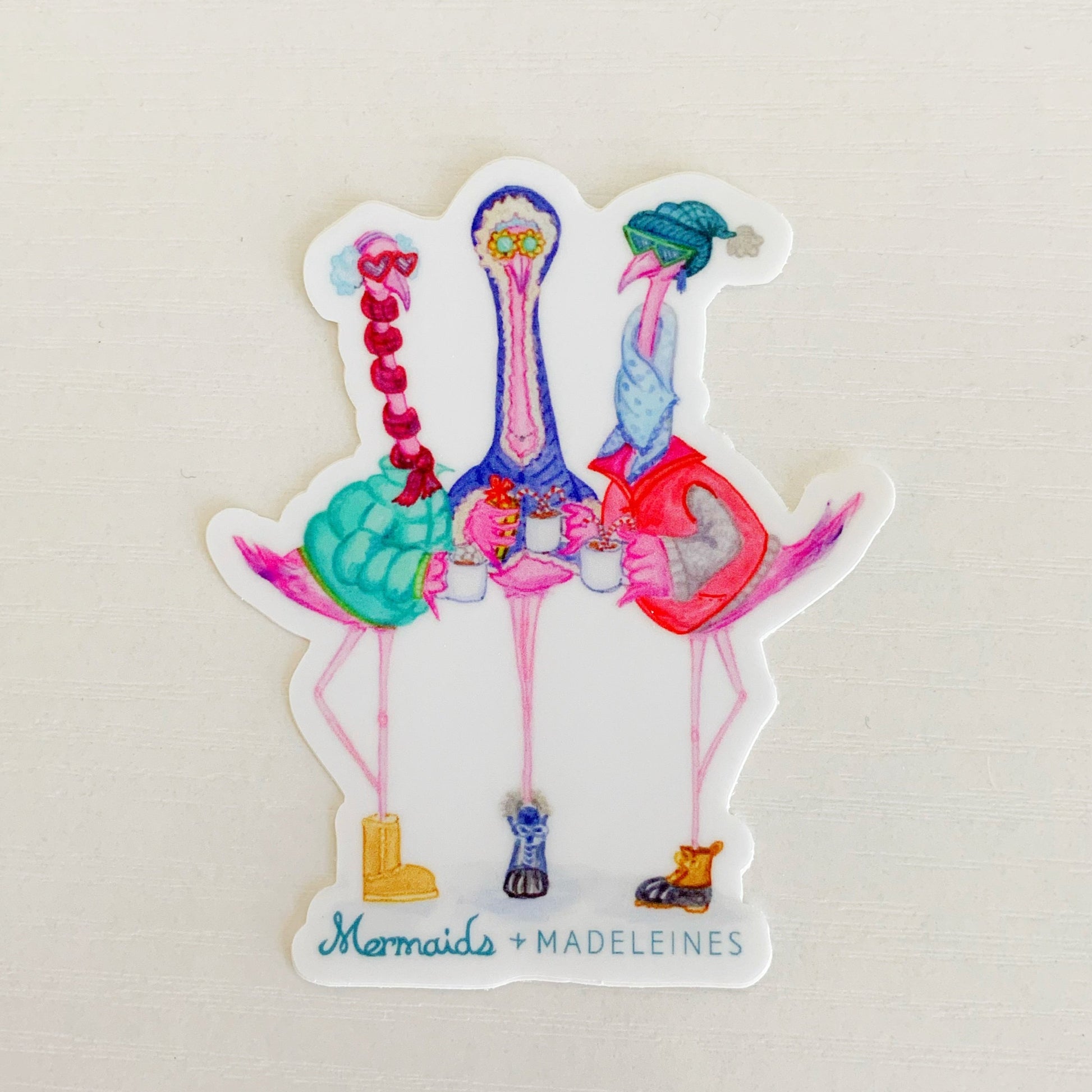 this is a photograph of the Flamingal winter vinyl sticker. the sticker is an illustration of 3 flamingos dressed in winter wear. This sticker is photographed on white surface