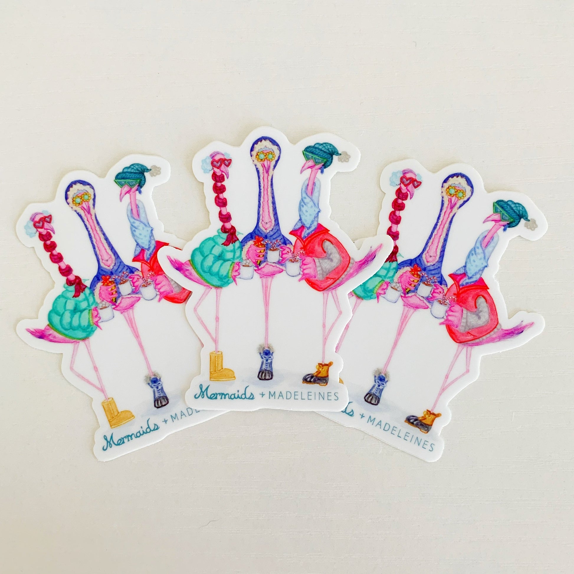 3 pack of winter flamingal vinyl stickers. the stickers are all the same. the sticker is an illustration of 3 flamingos dressed in winter wear, the artist is Heather Auclair Welch. this set of 3 stickers is photographed on a white surface