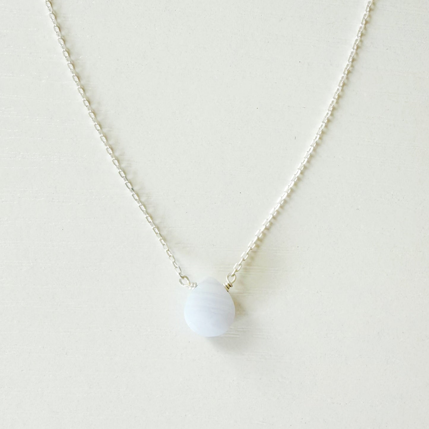A delicate silver chain necklace with a small, smooth, pale blue-white oval pendant, displayed against a clean, off-white background.