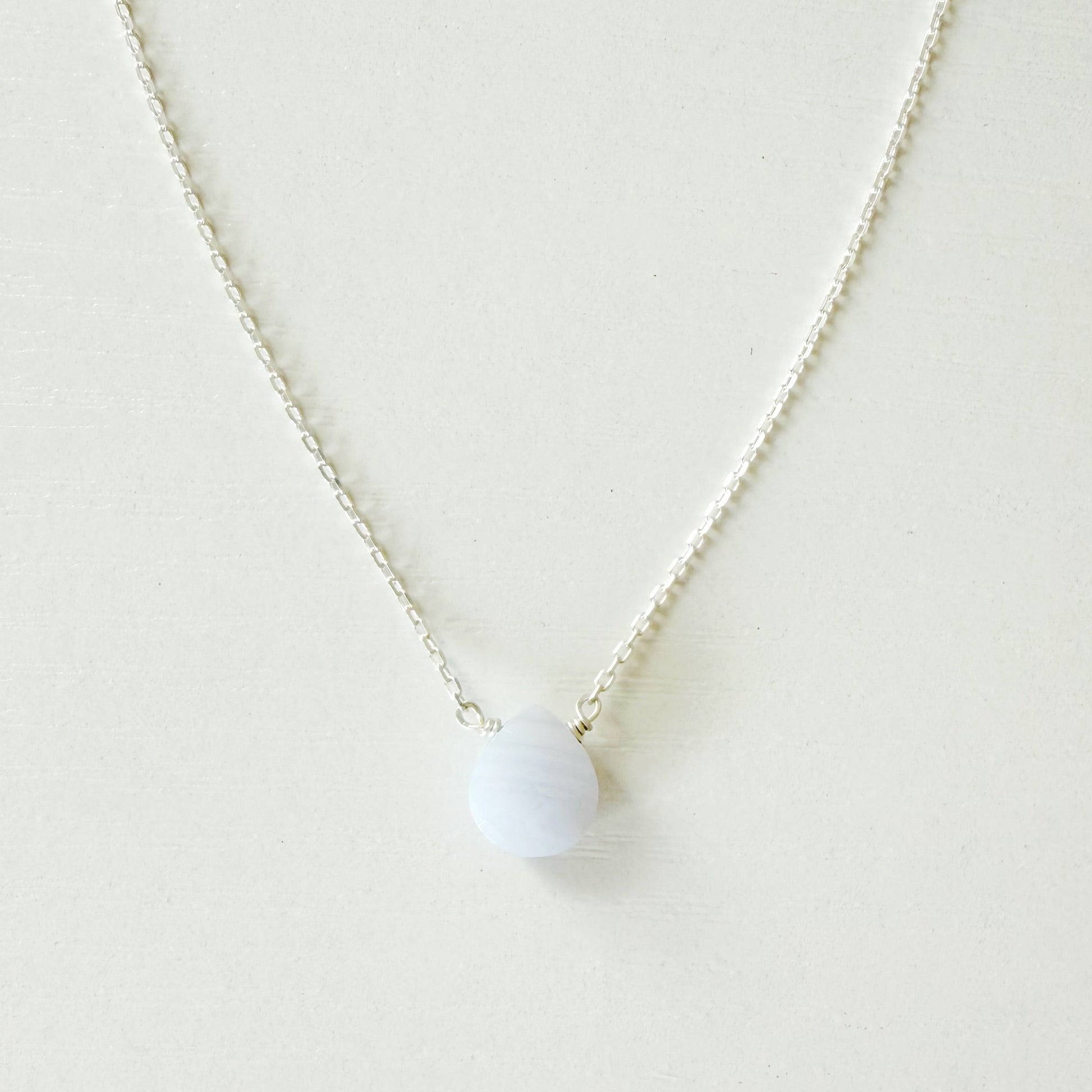 A delicate silver chain necklace with a small, smooth, pale blue-white oval pendant, displayed against a clean, off-white background.