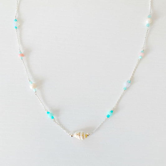 the island hopper necklace by mermaids and madeleines is a station style necklace with a shell at the center and semiprecious beads going up the sterling silver chain. this necklace is photographed flat and close up on a white surface