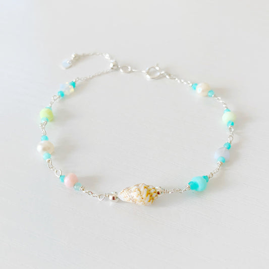 the island hopper adjustable bracelet by mermaids and madeleines is a station style bracelet with a shell at the center and semiprecious beads going up the sterling chain to a slide bead near the clasp. this bracelet is photographed close up with a front view on a white surface