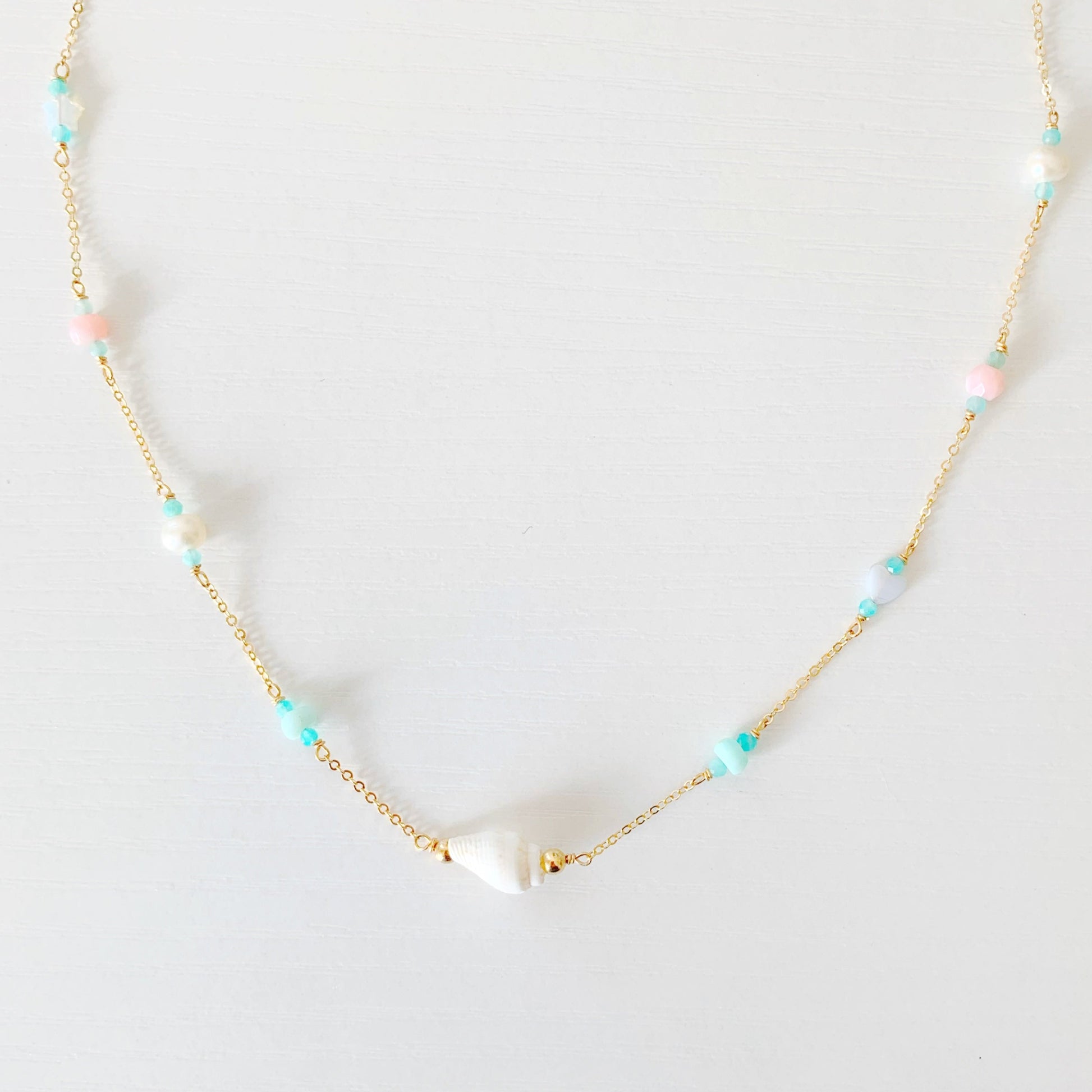 the island hopper necklace by mermaids and madeleines is a station style necklace with a shell at the center and semiprecious beads going up the 14k gold filled chain. this necklace is photographed close up on a white surface