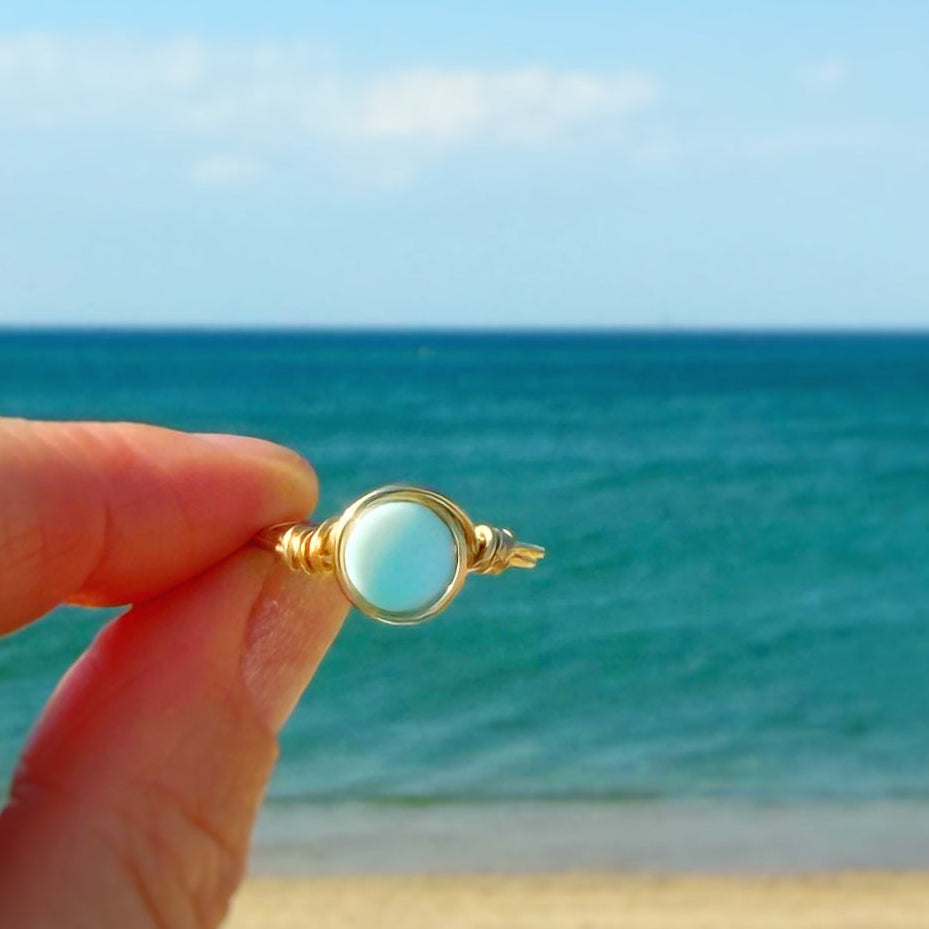 Gold filled wire-wrapped ring with a aqua Peruvian opal gemstone at the center held in a hand with a beach background