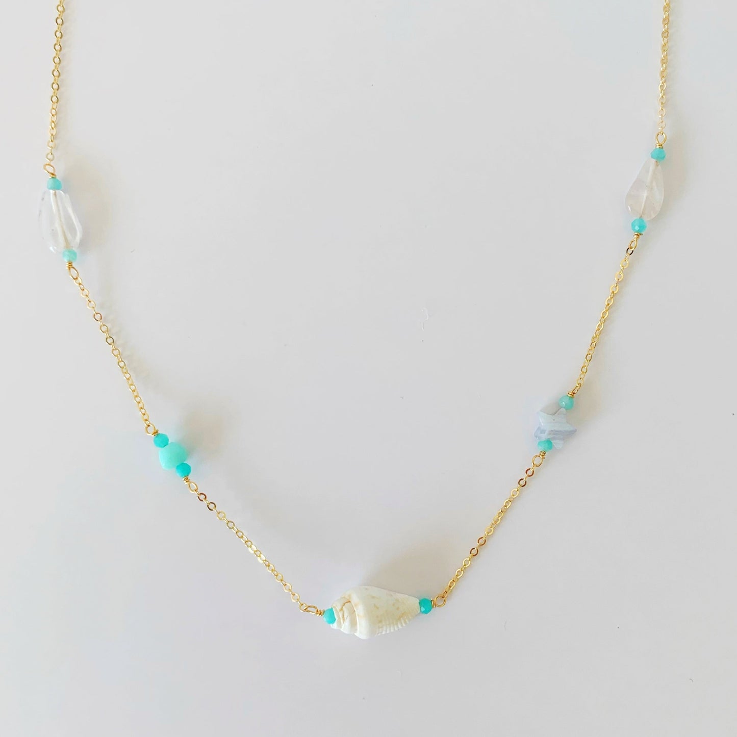 island hopper necklace by mermaids and madeleines is a station necklace with a shell at the center and a variety of semiprecious beads along the 14k gold filled chain to the clasp. this necklace is photographed on a white table