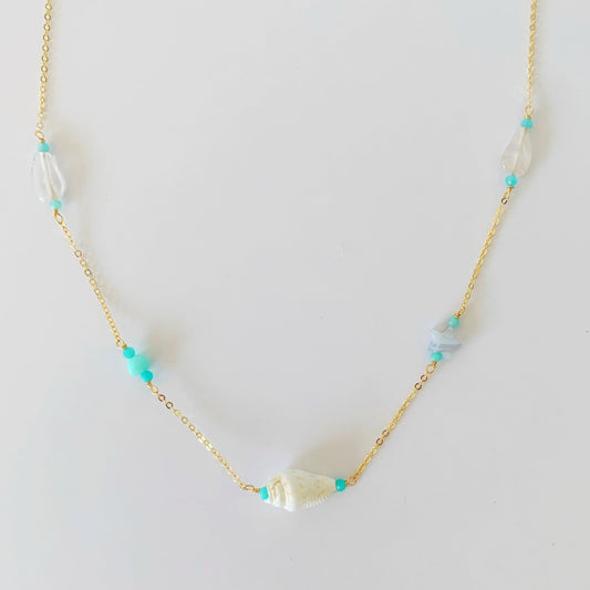 island hopper necklace by mermaids and madeleines is a station necklace with a shell at the center and a variety of semiprecious beads along the 14k gold filled chain to the clasp. this necklace is photographed on a white table