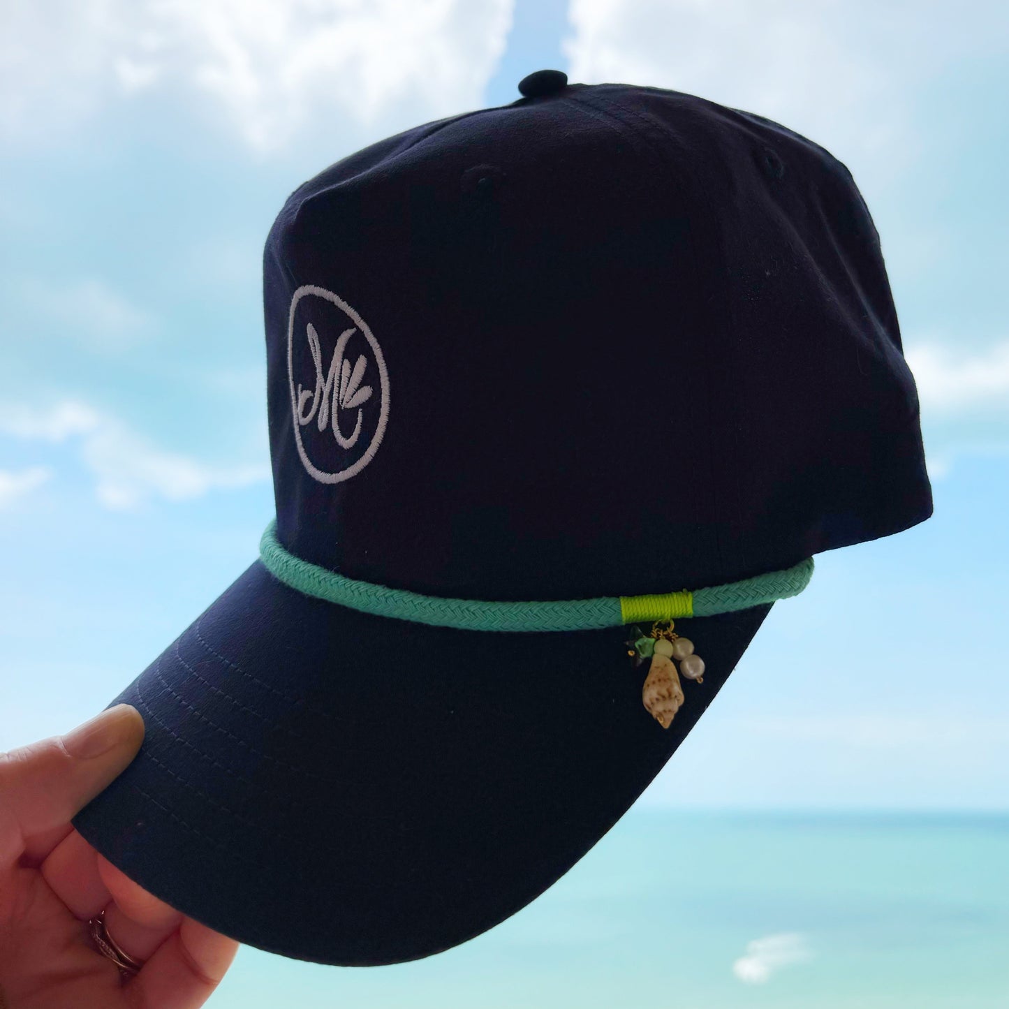 a navy blue snapback cap with a white "m' logo and teal rope around the bill held in a hand against a blue sky