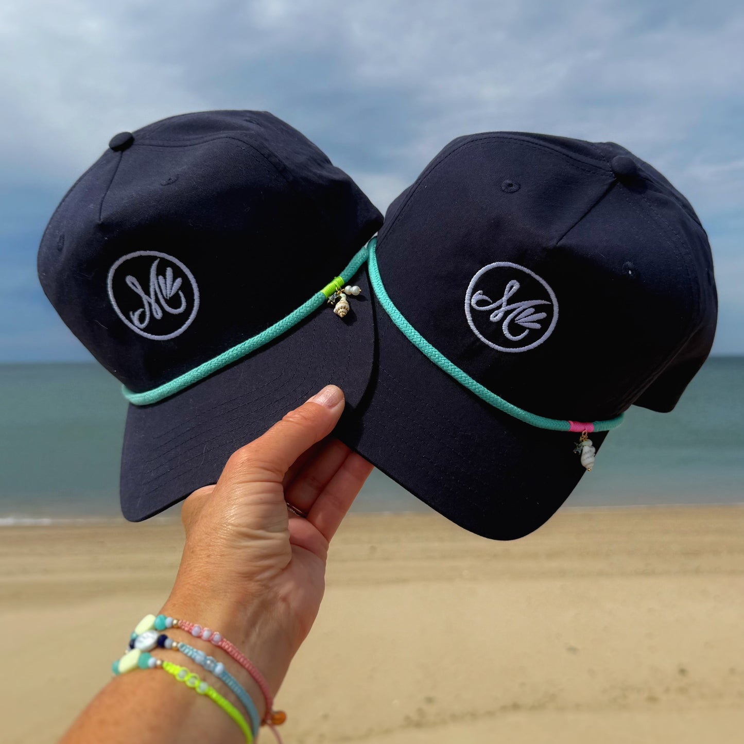 Two navy blue caps with a logo held by a hand on a beach.