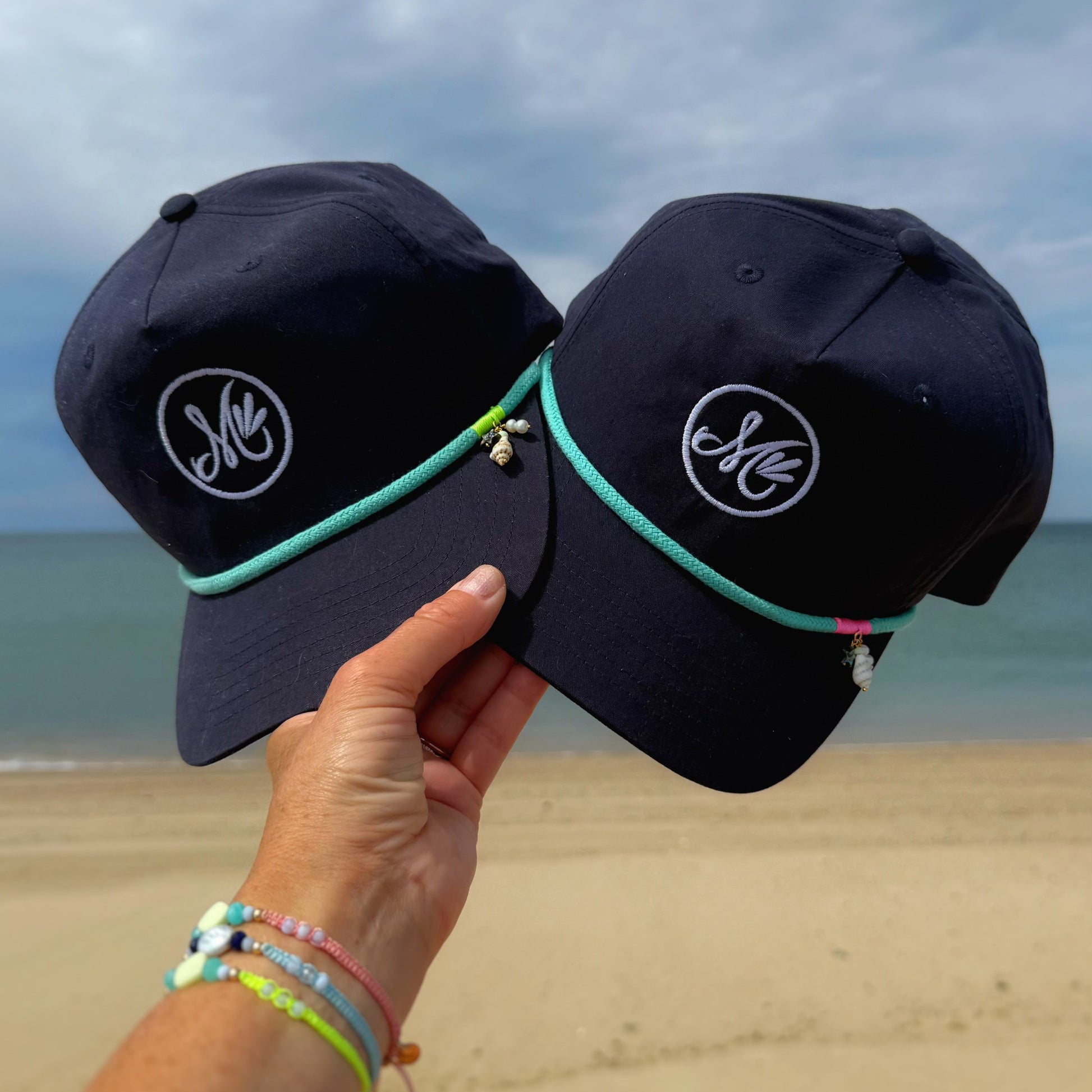Two navy blue caps with a logo held by a hand on a beach.