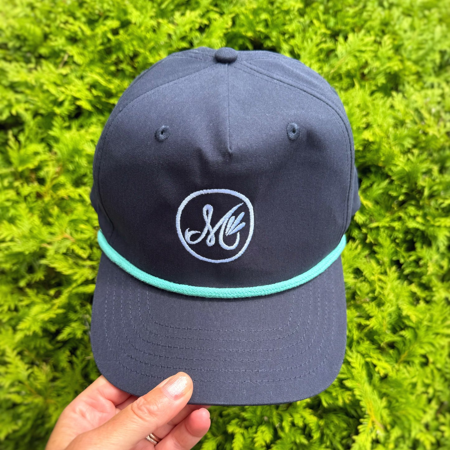 Navy blue cap with an M logo held in front of green tree